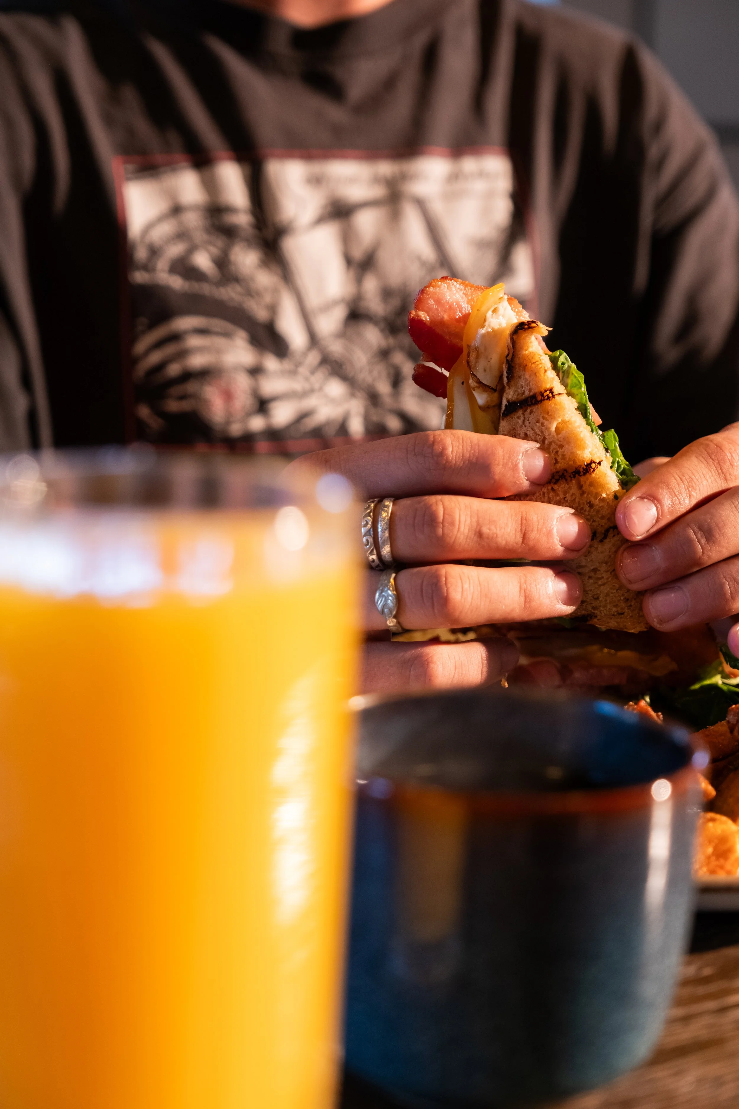 Person holding a grilled sandwich with bacon, cheese, lettuce, and tomato at a table, with a glass of orange juice and a small bowl in the foreground.