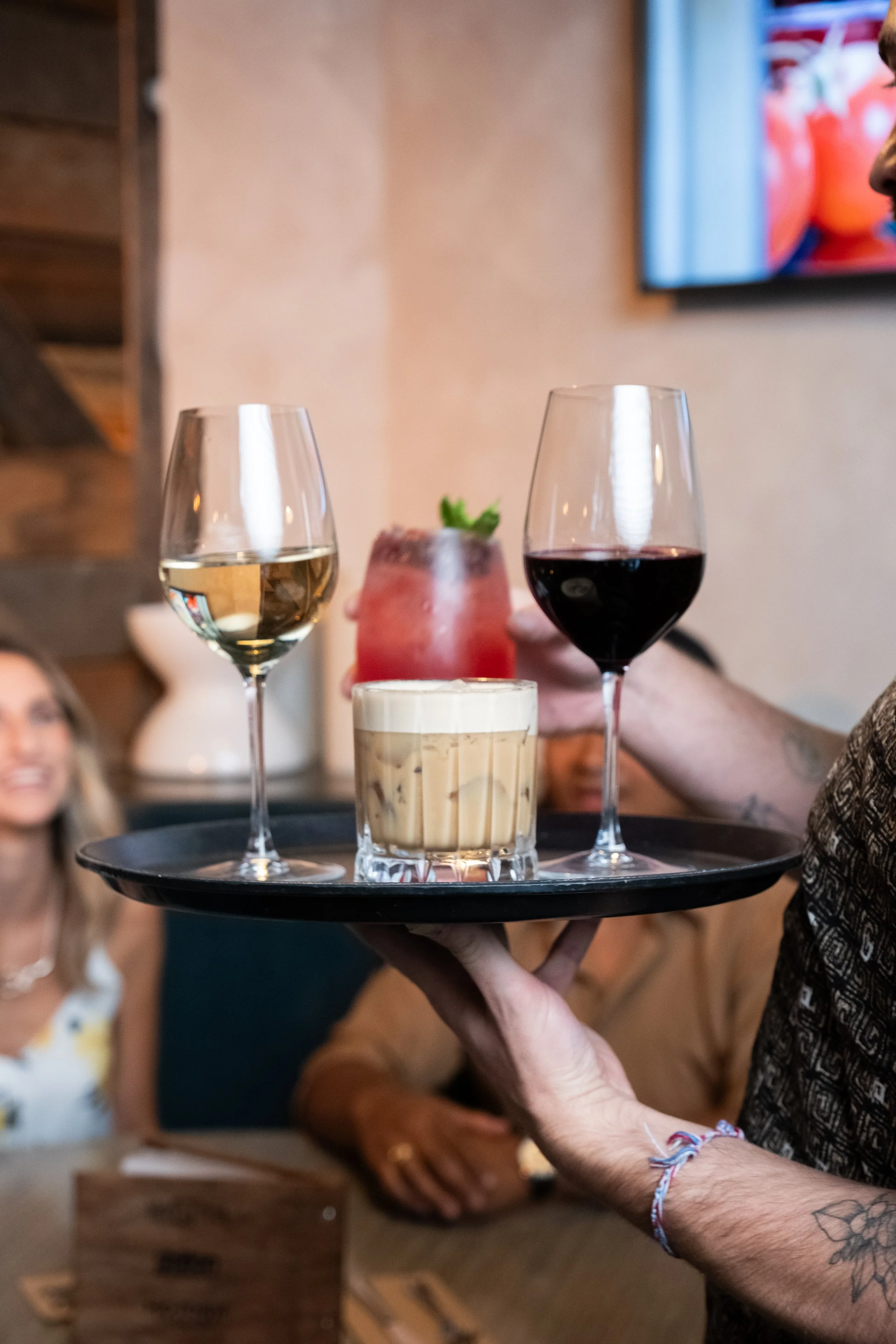 A server holds a tray with two glasses of white and red wine, a glass of a layered cocktail with a mint garnish, and a small container of sauce or dressing in a restaurant setting with people in the background.