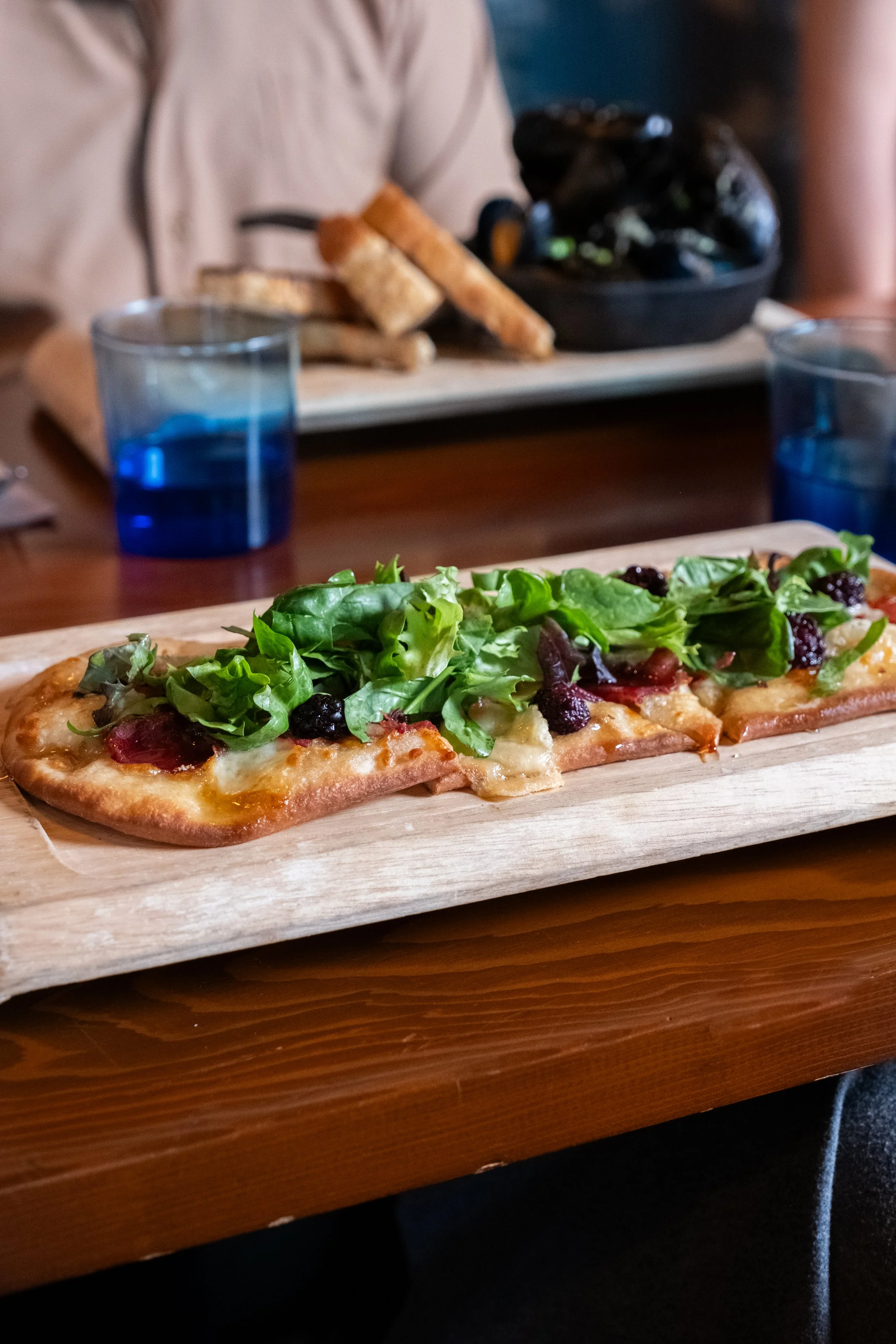 A rectangular pizza with arugula, blackberries, and melted cheese on a wooden serving board.