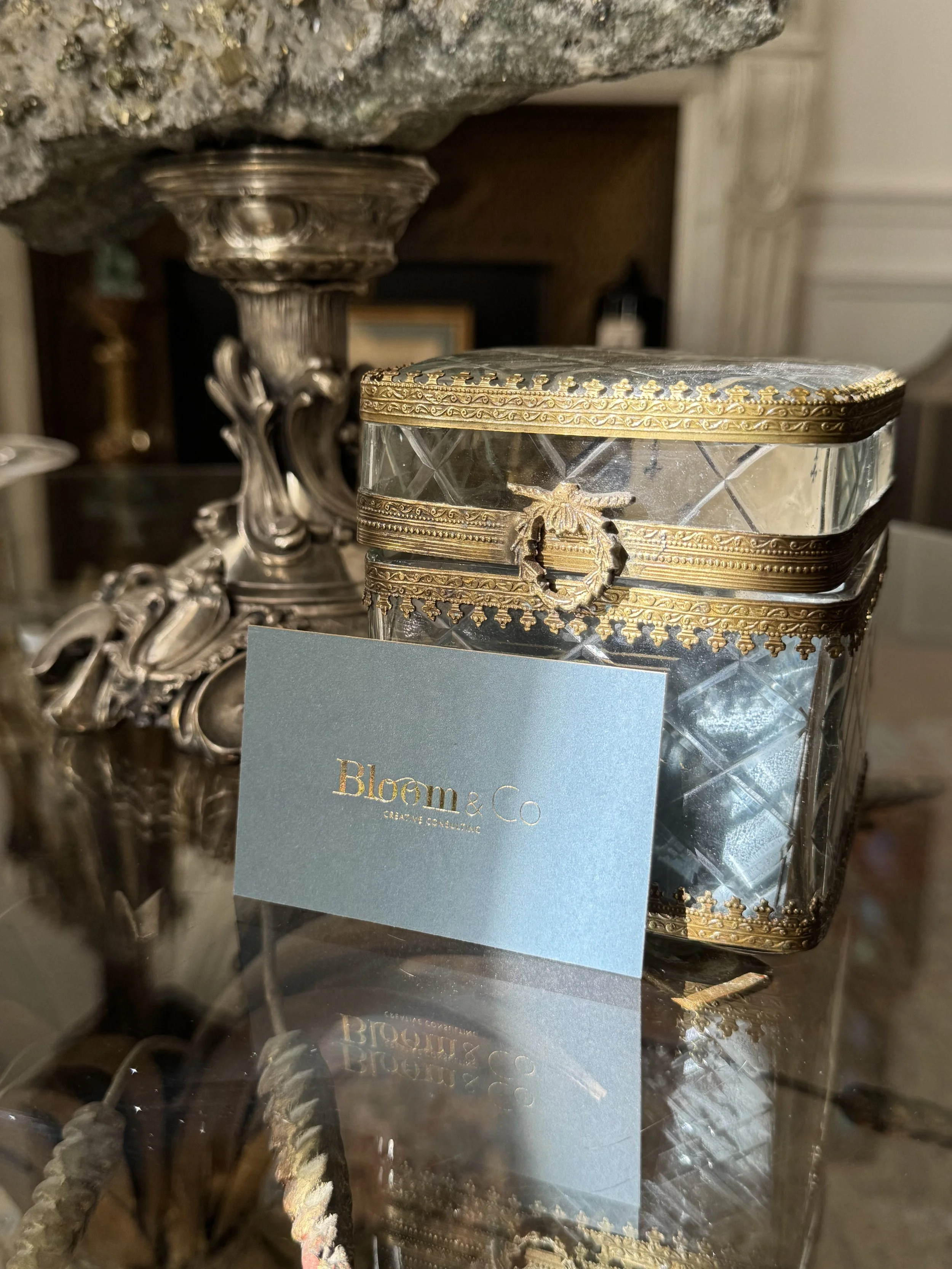 Une boîte en verre ornée de détails dorés, posée sur une surface réfléchissante, avec une carte portant l'inscription 'Bloom & Co'. En arrière-plan, un élément décoratif en métal argenté et une surface en pierre ou en bois.