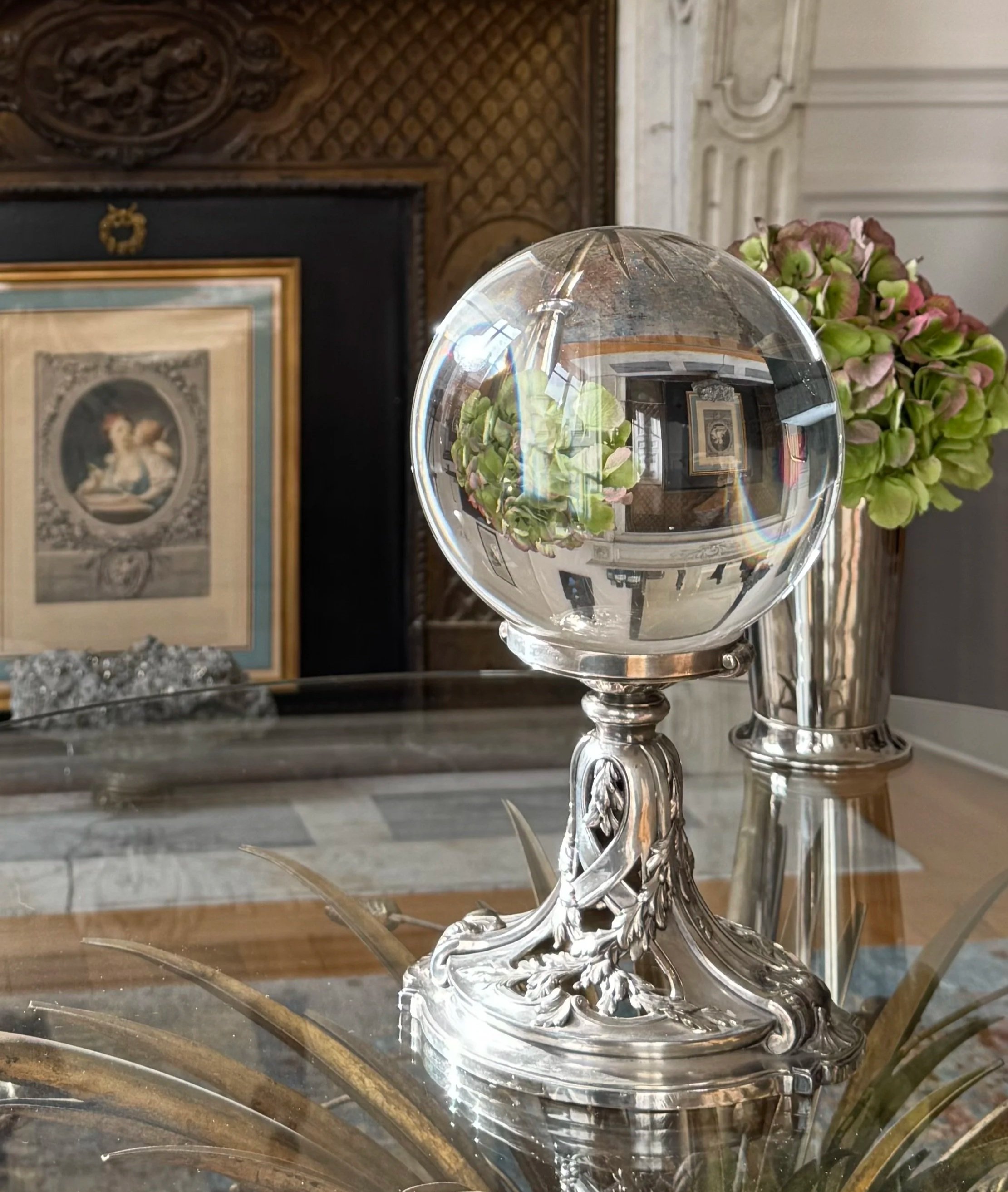Un cristal transparent en forme de boule placé sur un support en métal orné de détails floraux, à côté de fleurs vertes et roses, dans un cadre d'intérieur avec des murs en pierre et des œuvres d'art encadrées.