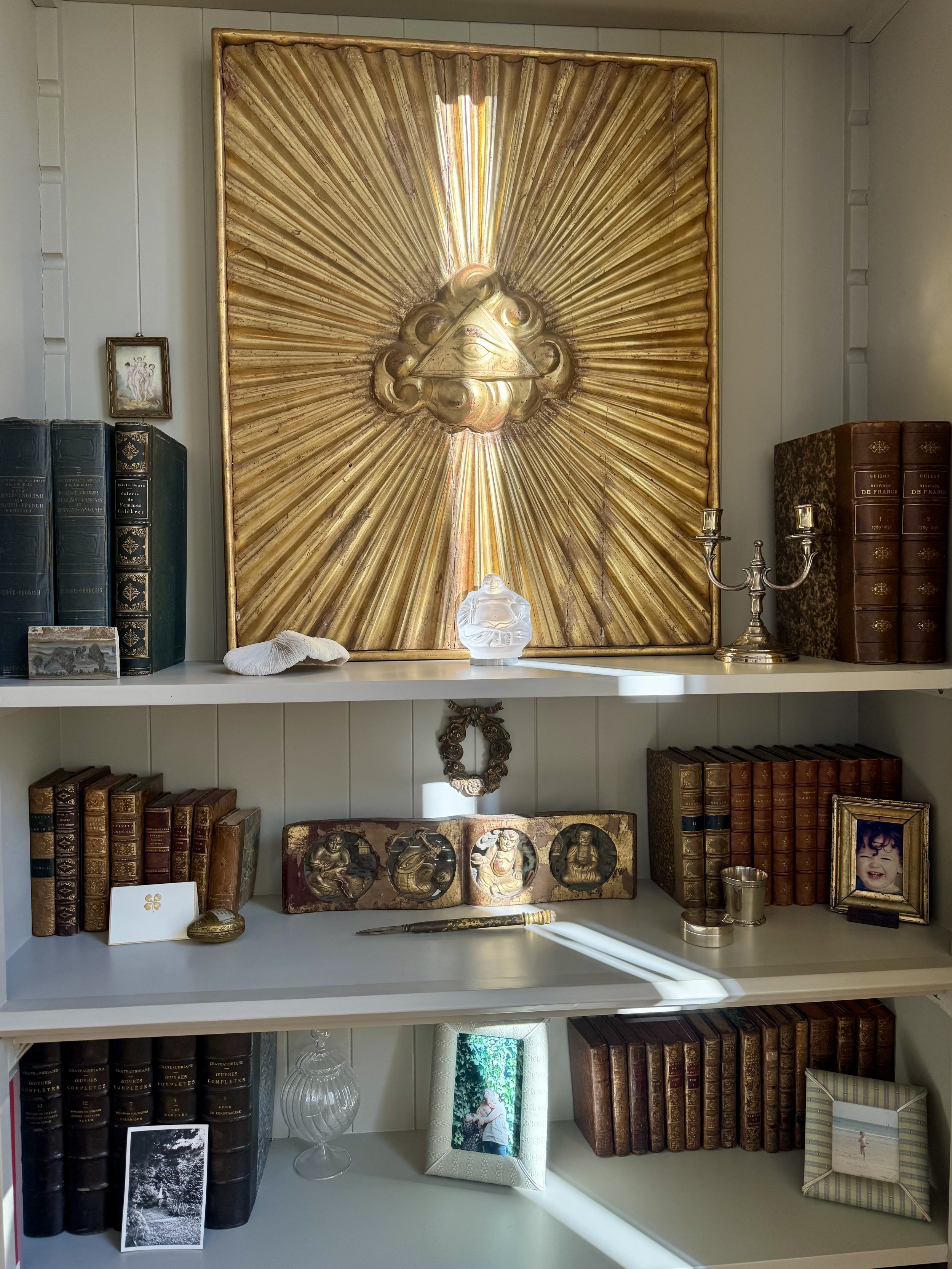 Une bibliothèque avec des livres anciens, une croix en or, une photo de famille, une statuette, un cadre avec une photo d'enfant, un miroir et divers objets décoratifs.