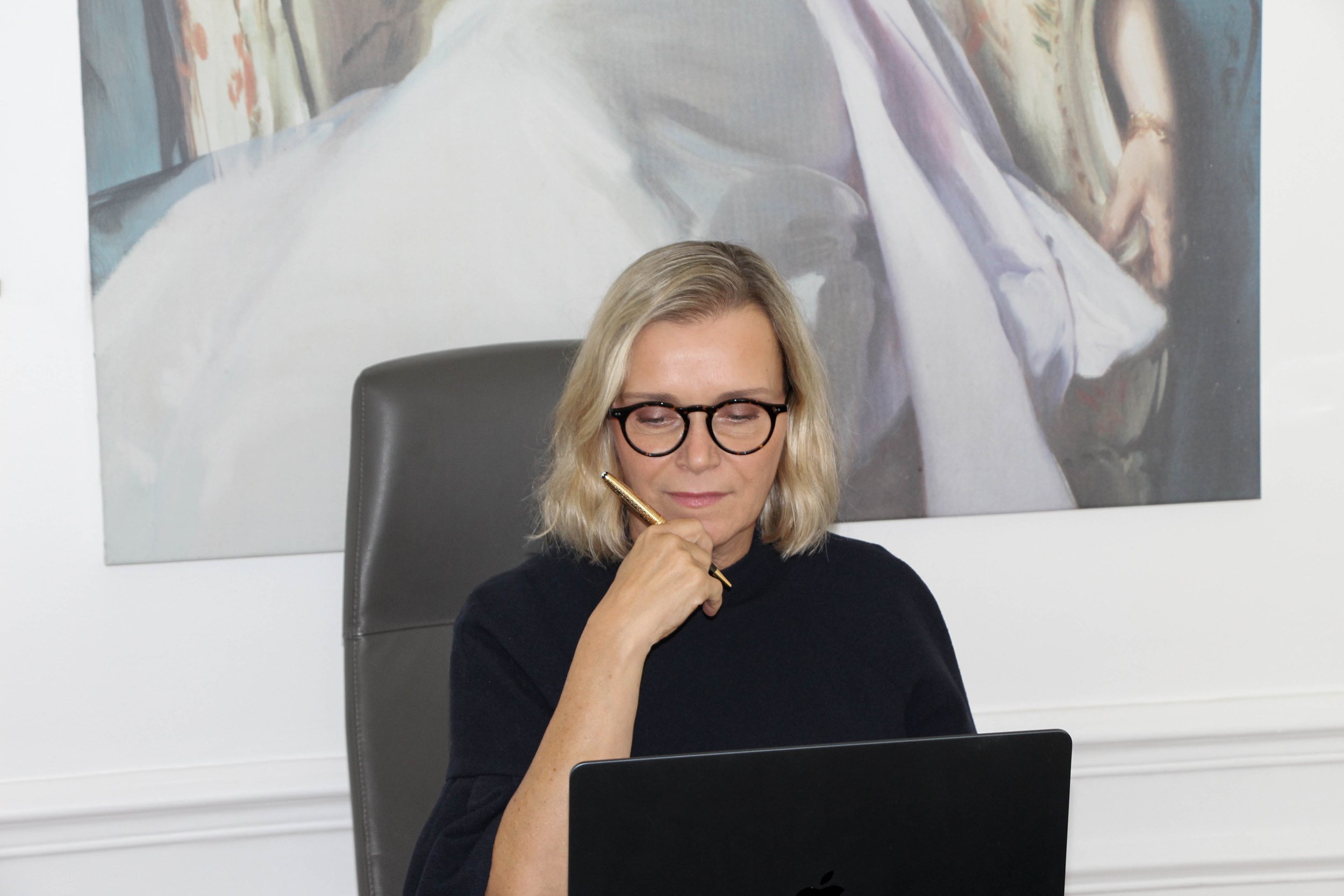Une femme aux cheveux blonds, portant des lunettes rondes noirs, assise à un bureau, en train de réfléchir ou de travailler, avec un stylo doré à la main. Derrière elle, un tableau ou une grande image d'une scène artistique ou religieuse.