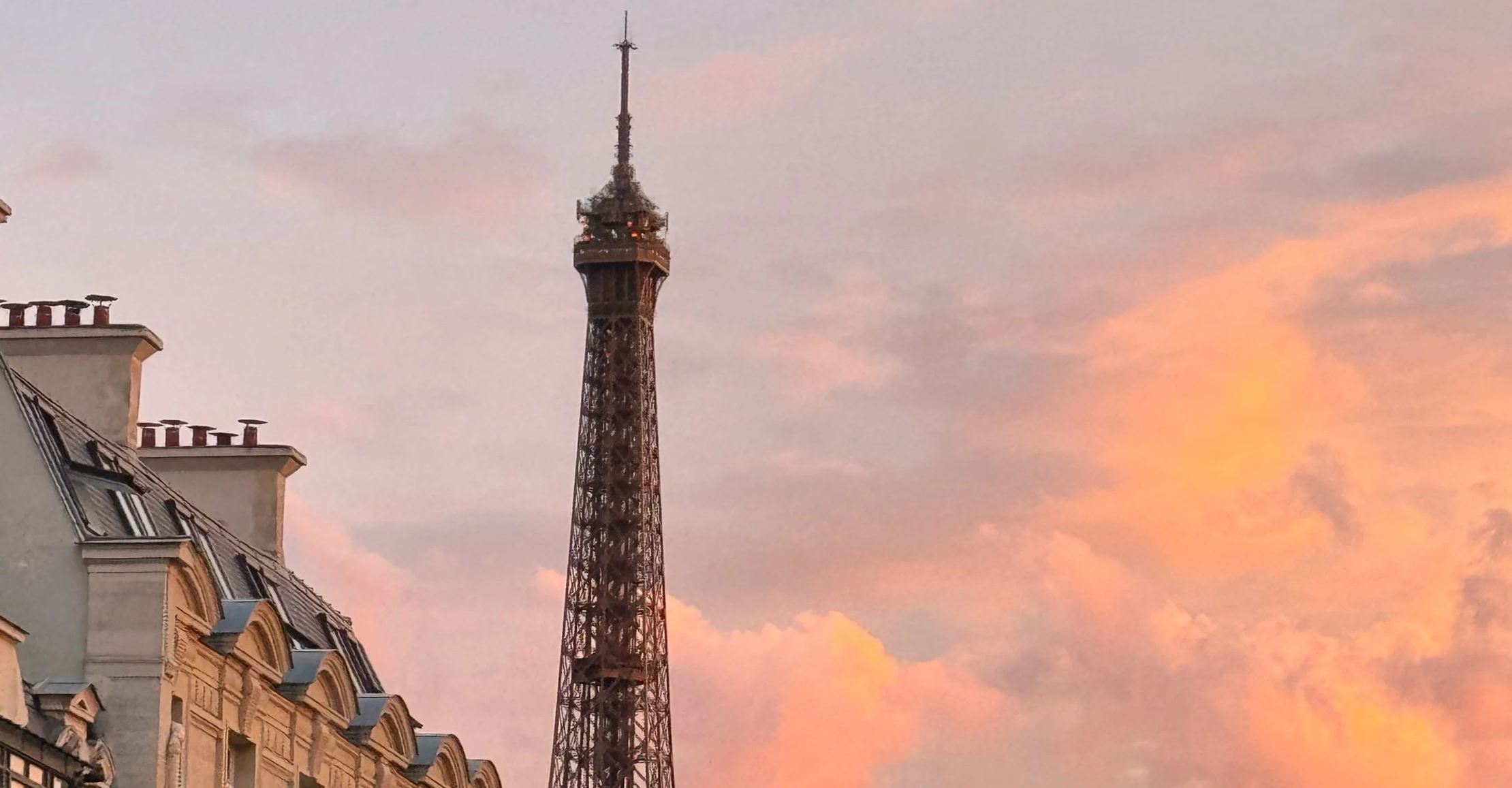 La Tour Eiffel, visible derrière des immeubles haussmaniens, sous un ciel pastel au coucher du soleil.