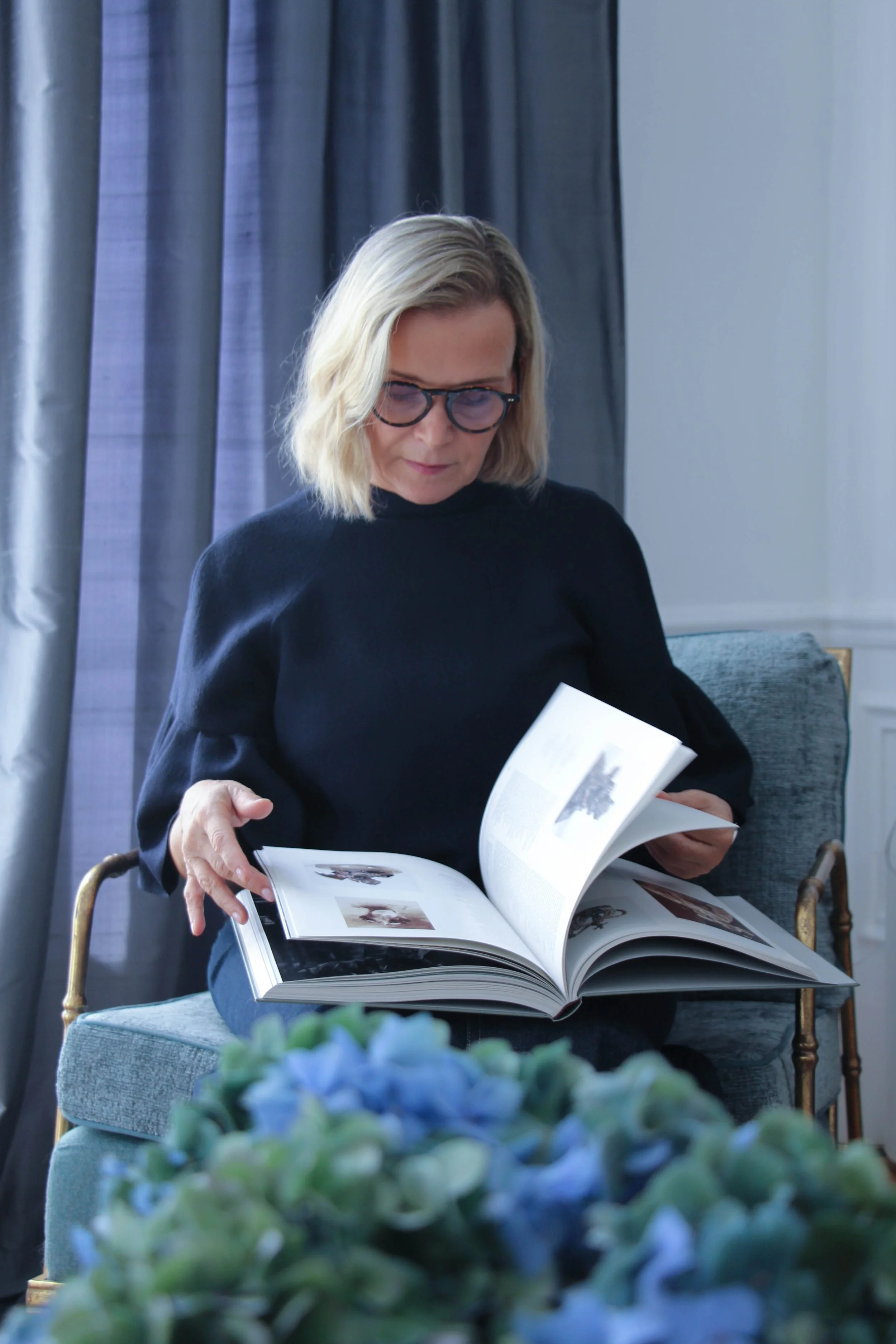 Une femme avec des lunettes, portant un pull noir, assise sur une chaise en bois, en train de feuilleter un livre illustré. En avant-plan, on voit un bouquet de fleurs bleues.