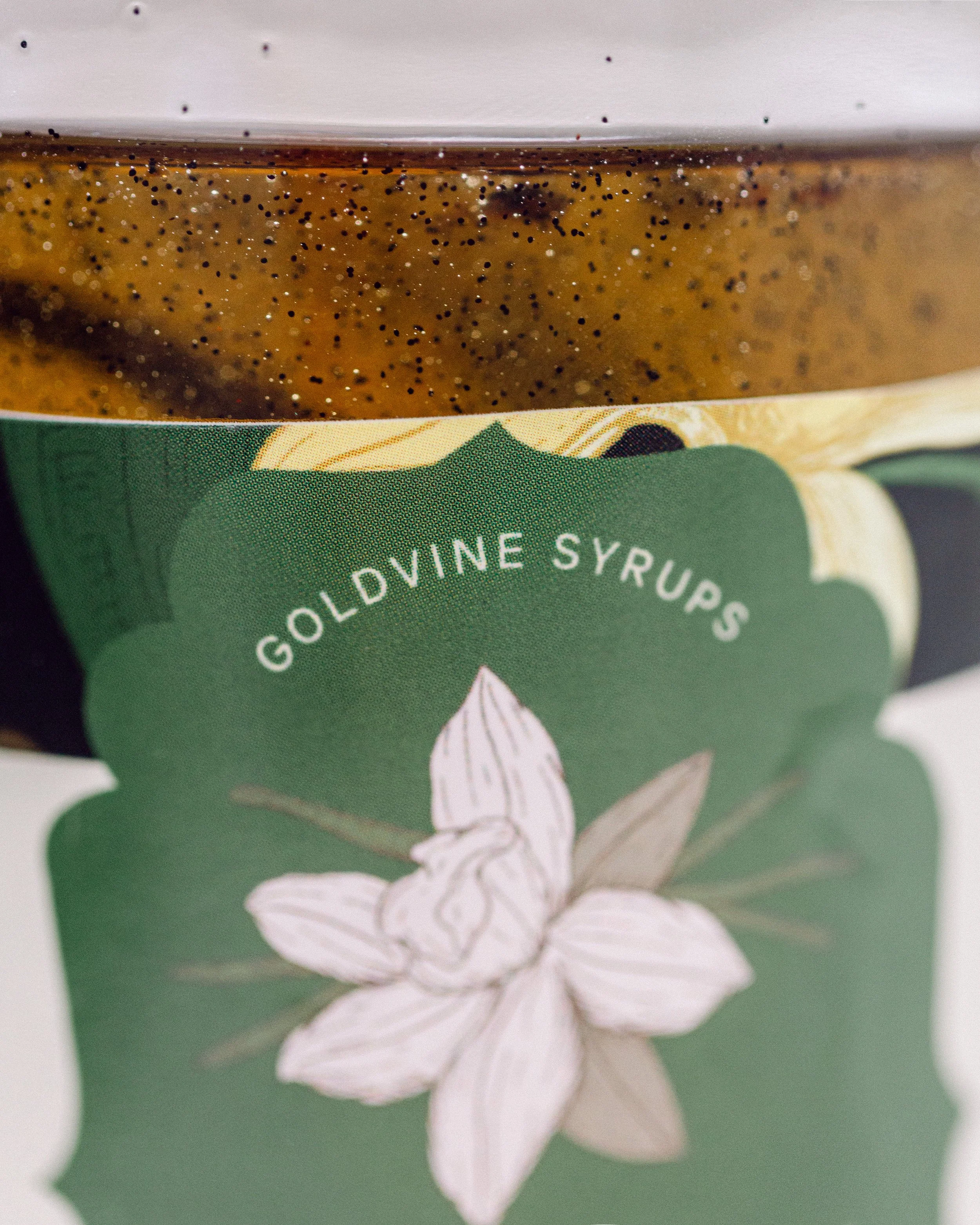Close-up of a jar of golden syrup with visible black and white specks, and a green label featuring an illustration of a white flower.
