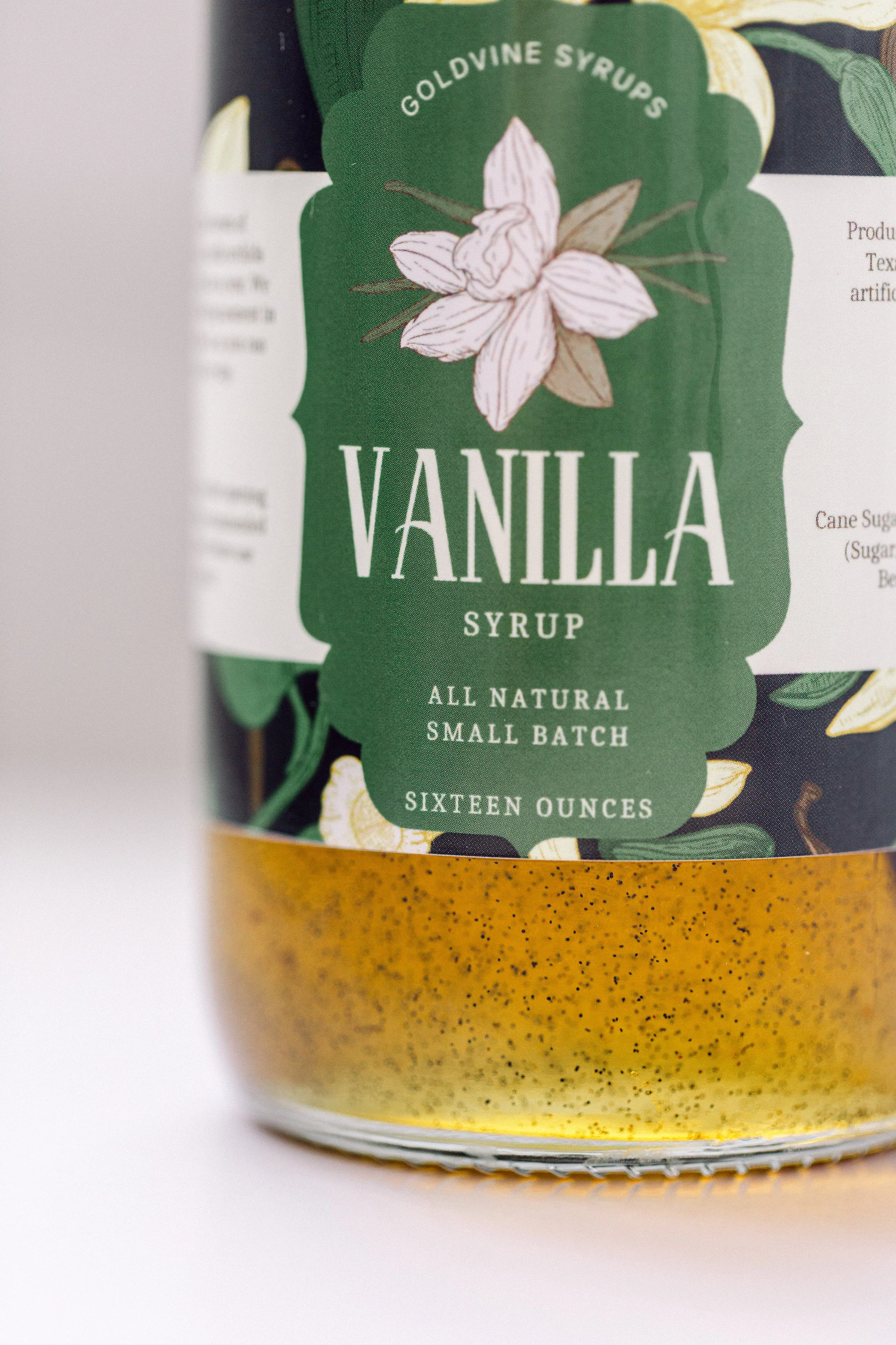 Close-up of a green label on a container of vanilla syrup from Golddivine Syrups. The label features an illustration of vanilla flowers and leaves, and text indicating it is all natural, small batch, with a net weight of sixteen ounces.