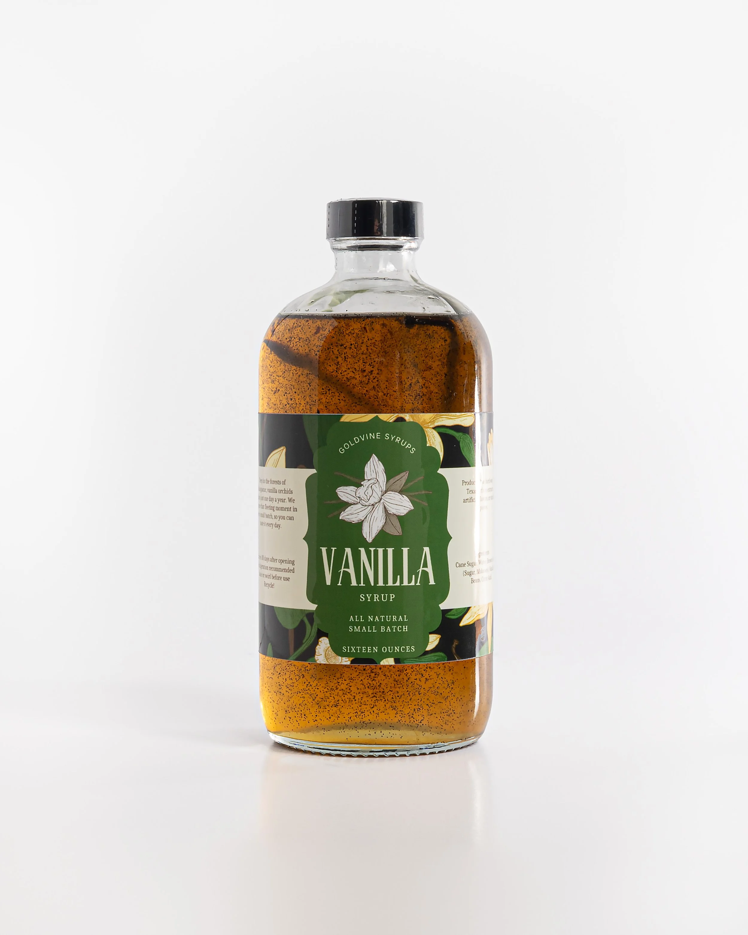 A clear glass bottle filled with vanilla syrup, featuring a white label with a green background, a vanilla flower illustration, and black cap, placed on a white surface against a white background.