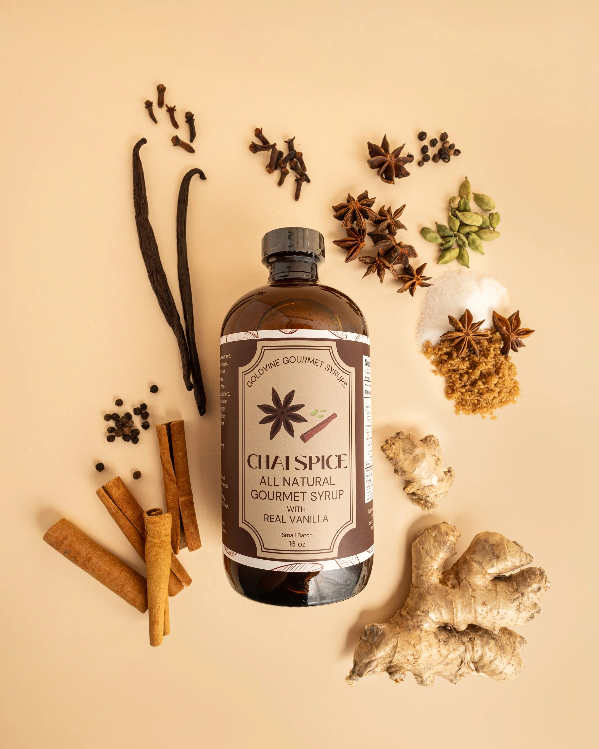 A bottle of Goldvine Gourmet Syrups chai spice with slices of cinnamon, star anise, black peppercorns, cardamom pods, ginger root, and pieces of dried ginger arranged around it on a beige background.
