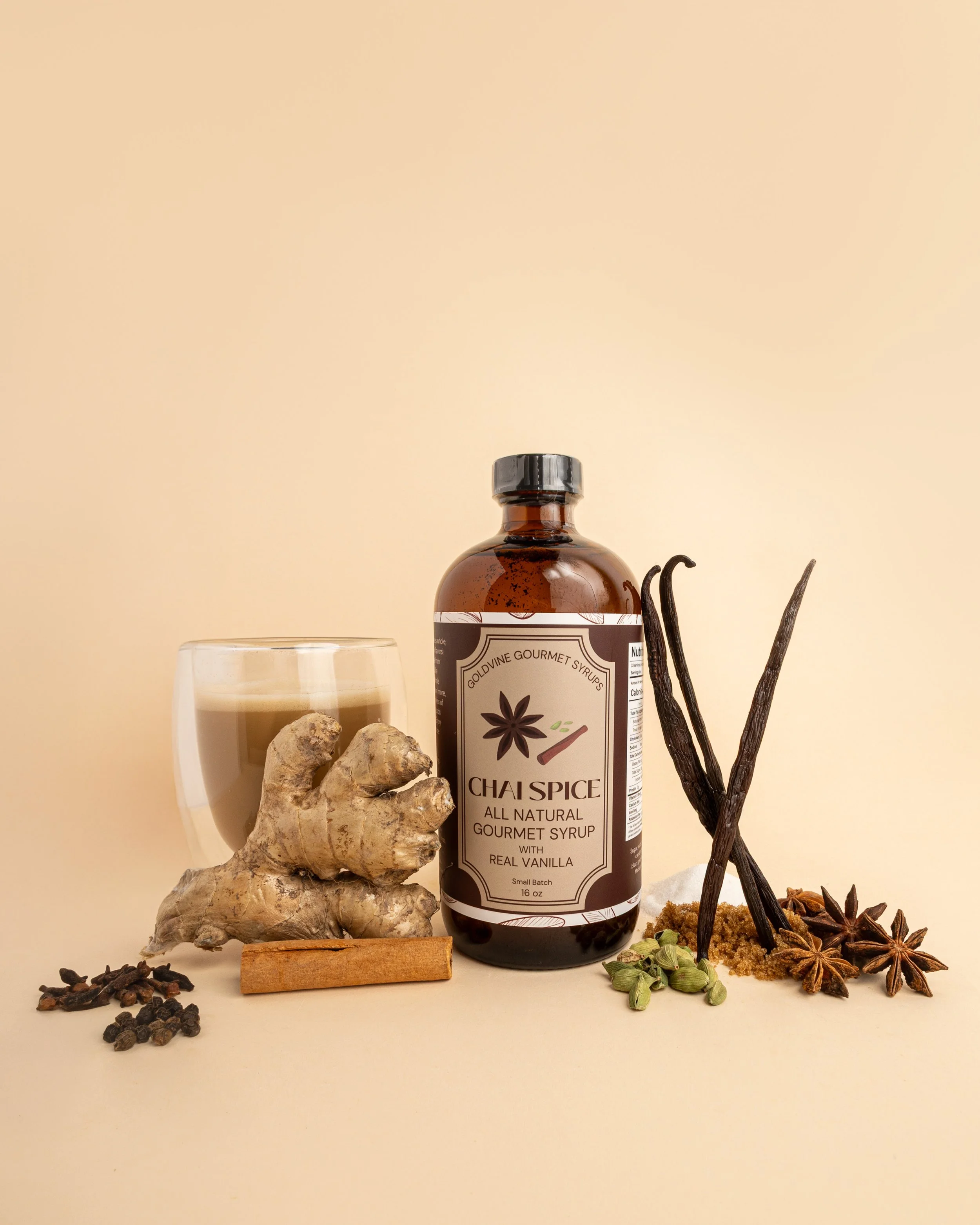 A bottle of chai spice syrup surrounded by spices including ginger root, star anise, cloves, green cardamom, black peppercorns, cinnamon stick, and vanilla beans, with a glass of chai latte