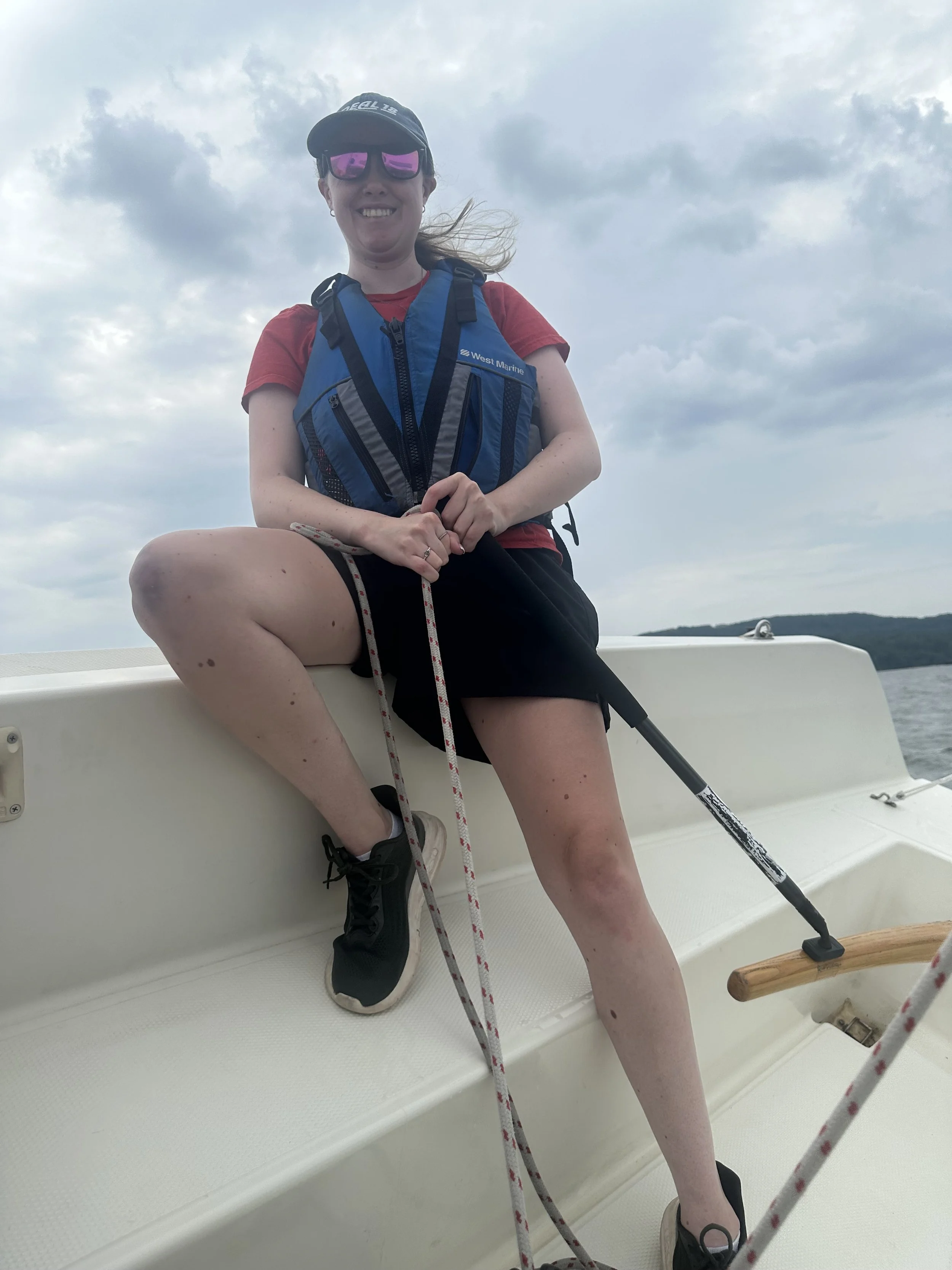 A smiling woman wearing sunglasses, a baseball cap, a red t-shirt, and a blue life vest, sitting on a boat with one leg propped up on the side, holding a rope, with water, hills, and a cloudy sky in the background.