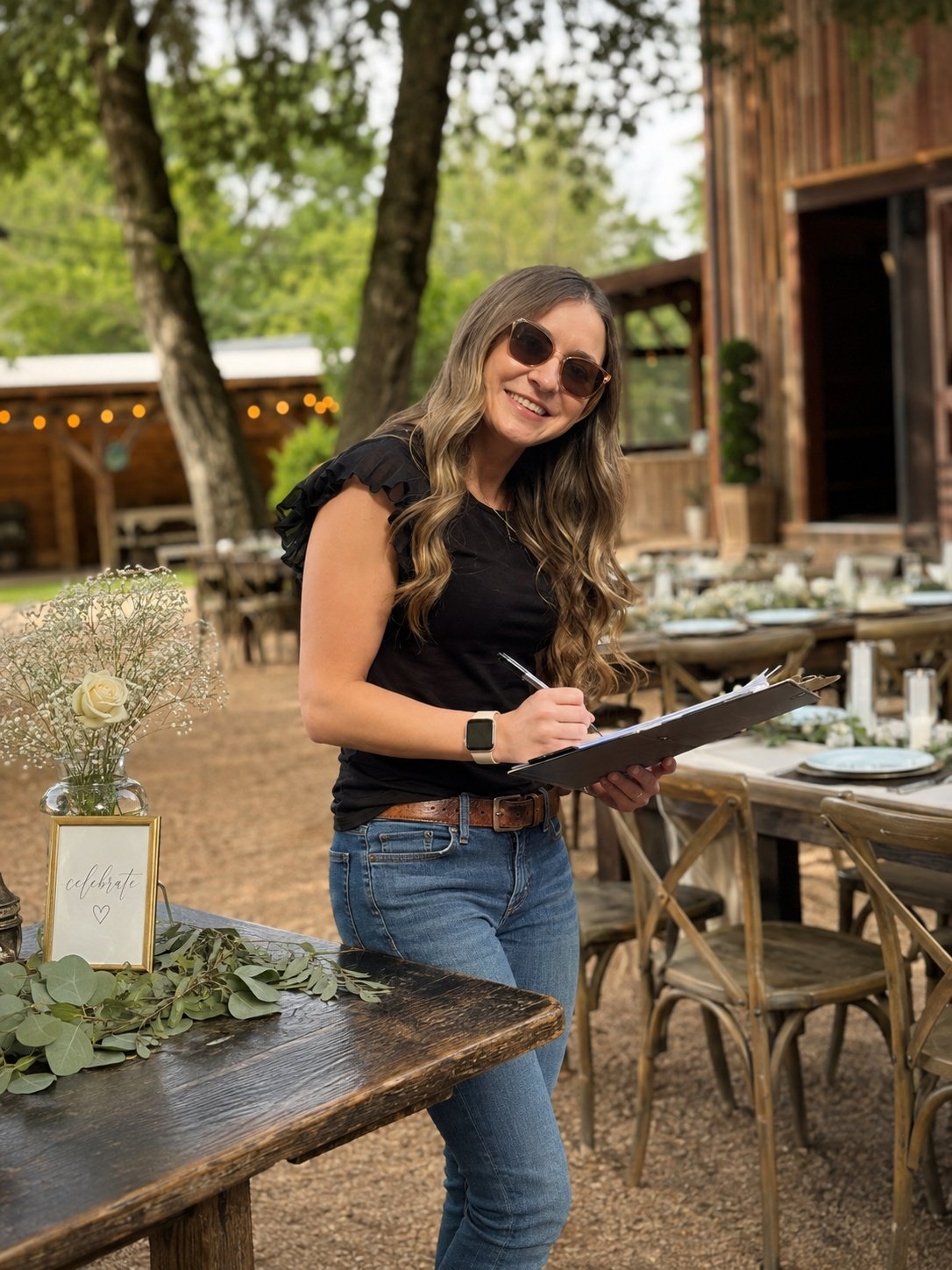A woman wearing sunglasses and a black shirt, smiling while writing on a clipboard outdoors at a rustic event setting with wooden tables and chairs, decorative greenery, and a sign that says 'celebrate' with a heart.
