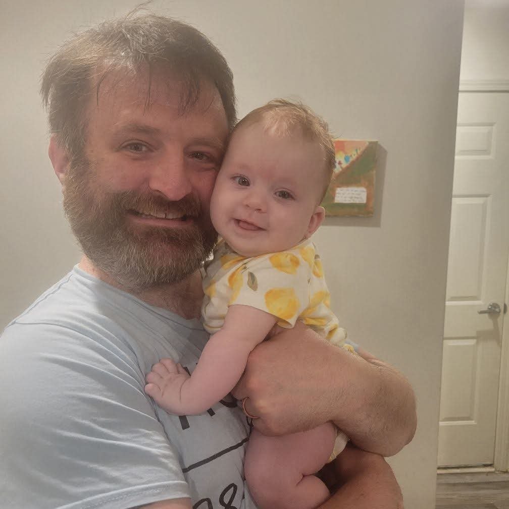 A man with a beard and a mustache holding a smiling baby girl with curly hair. The man has grayish hair and is wearing a light-colored T-shirt. The baby wears a yellow and white patterned dress. They are indoors, standing in front of a wall with a small colorful painting and a closed white door in the background.
