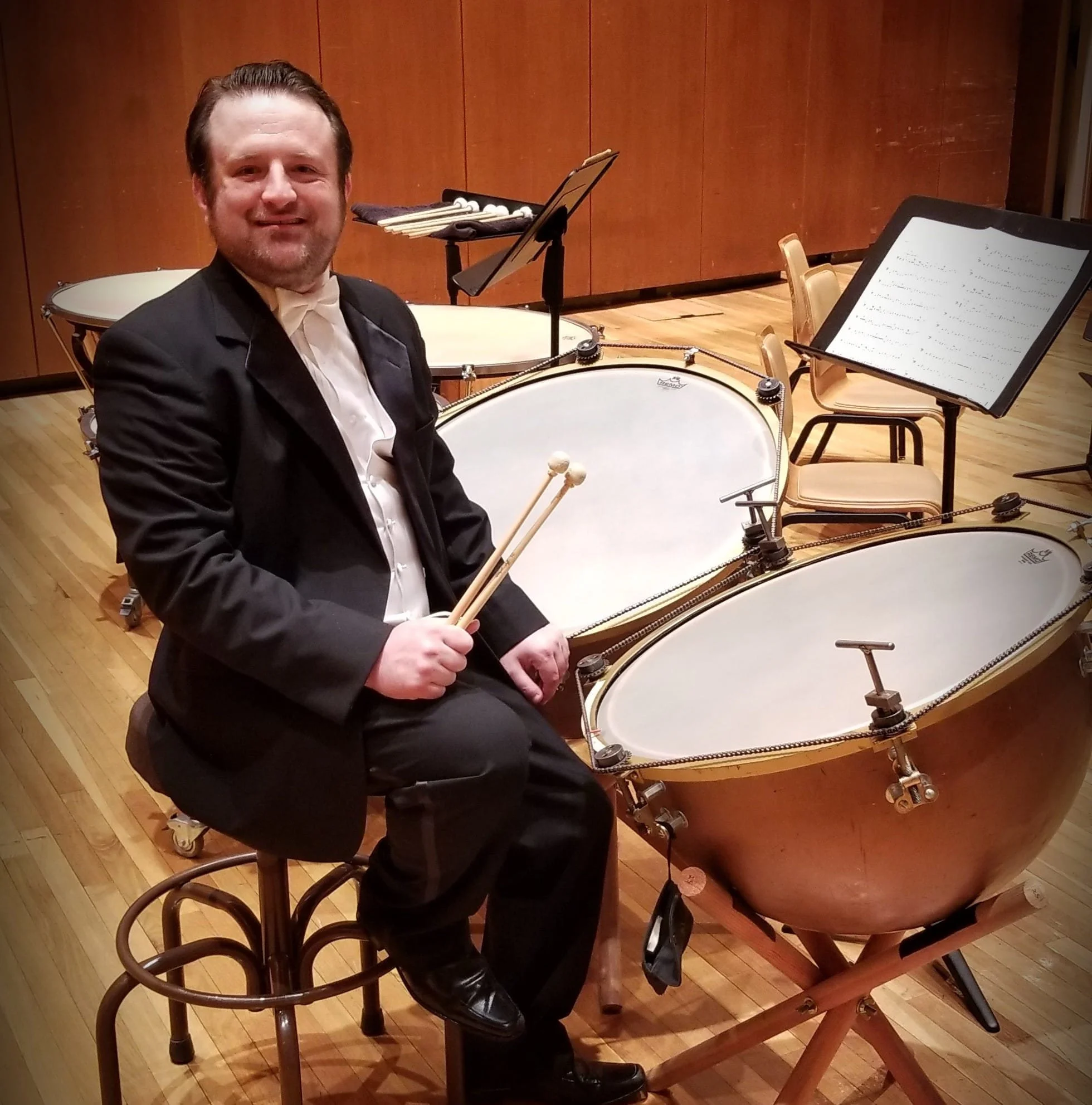 A man in a tuxedo holding drumsticks sitting near a timpani in a concert hall.