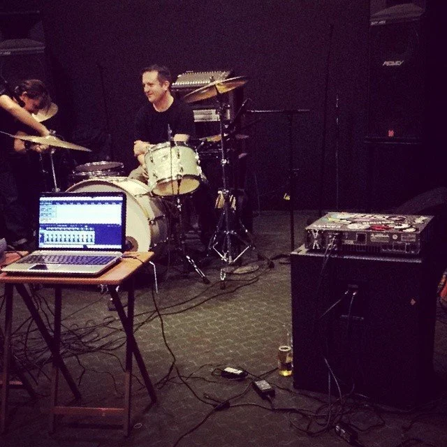 A man sitting behind a drum set with a laptop on a table in front of him, in a music rehearsal space with black walls and audio equipment.