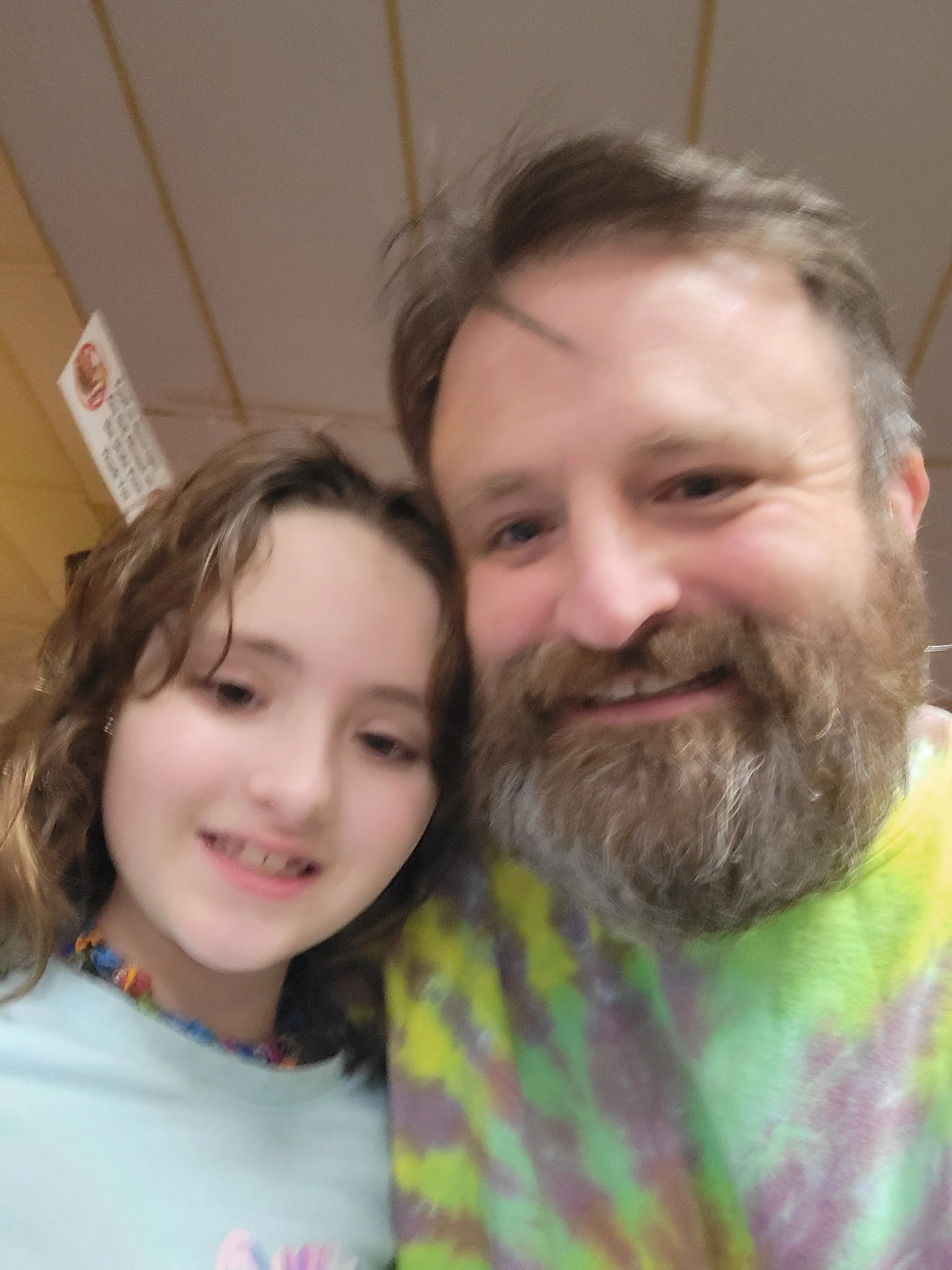 A young girl and a man smiling for a close-up selfie against a brown wooden paneled wall, with part of a colorful tie-dye shirt visible on the man.