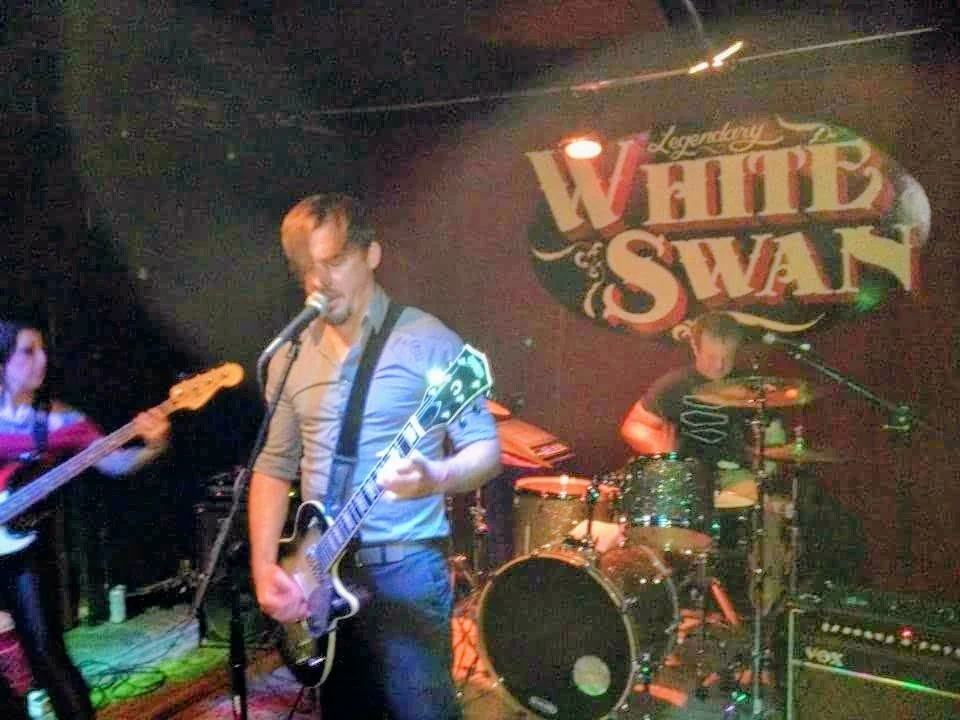 Musicians perform live on stage at White Swann venue, including a guitarist singing into a microphone and a drummer behind a drum set, with a dark backdrop and band logo.