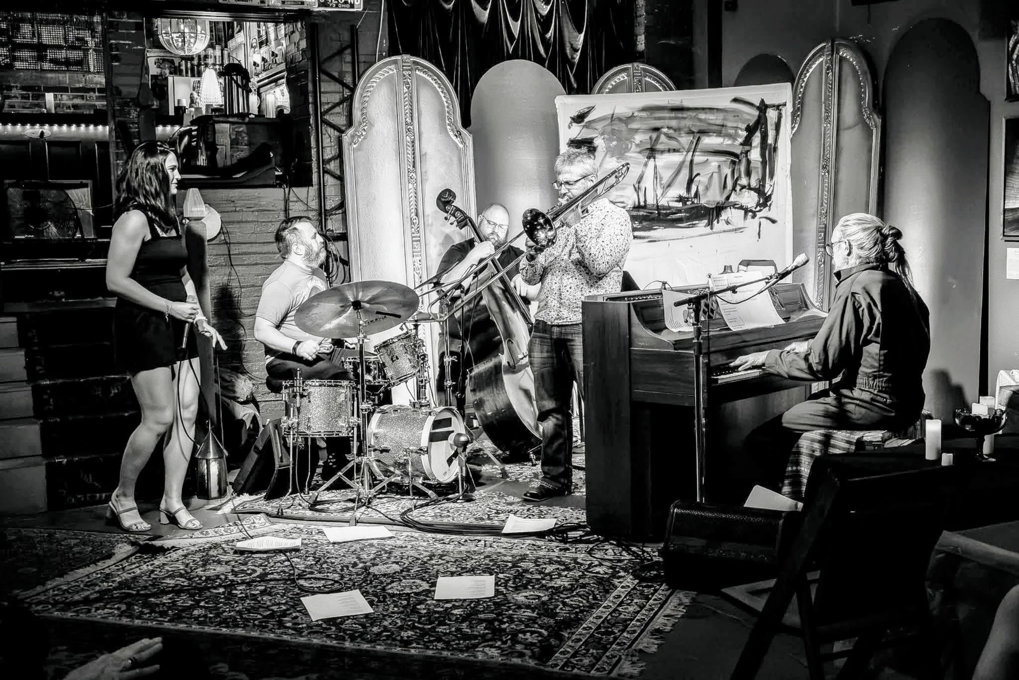 A jazz band performing on stage with a pianist, trombone player, and other musicians, in a room with ornate mirrors and paintings.