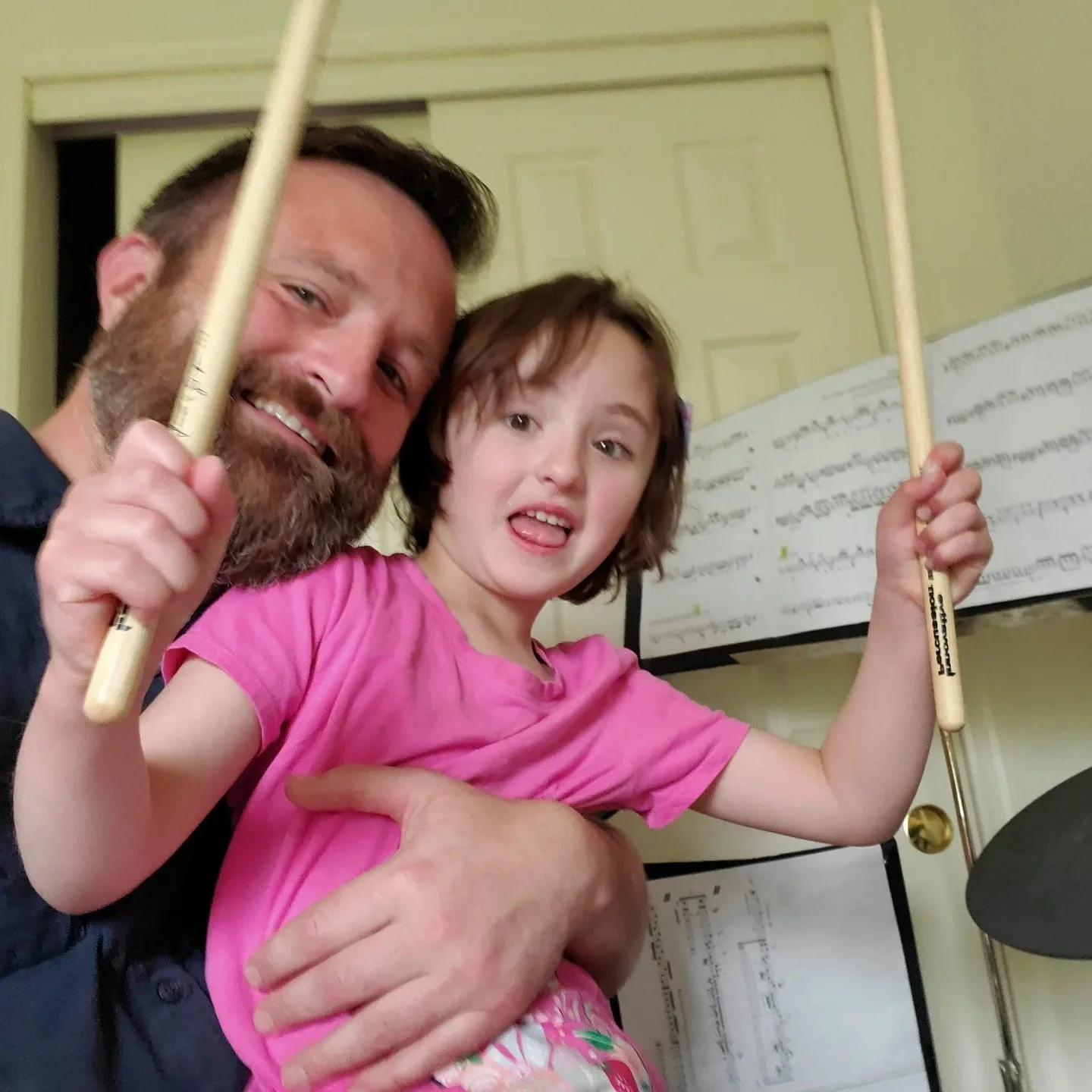A man and a young girl are smiling and holding drumsticks, standing in front of a music sheet with a closed door in the background.