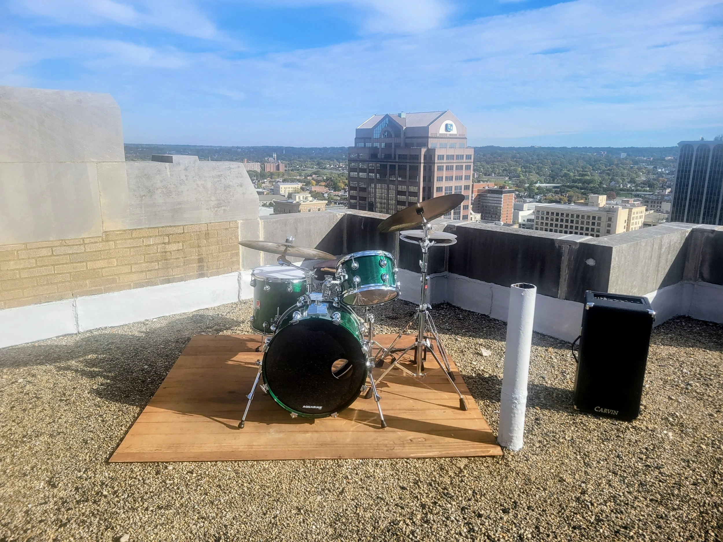 A rooftop with a drum set on a wooden platform, overlooking a city skyline with tall buildings and a blue sky with clouds.