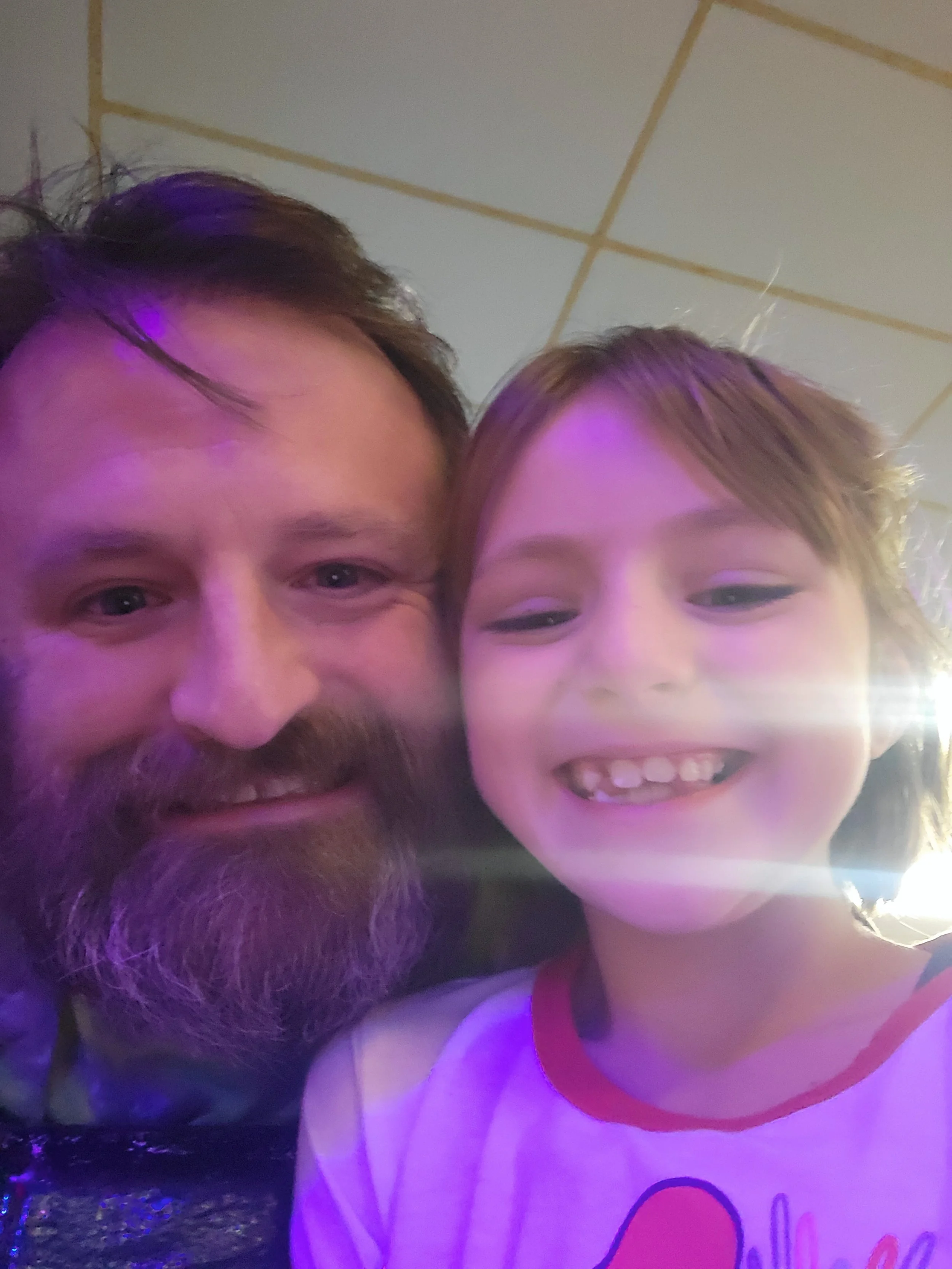 A close-up selfie of a man with a beard and a woman with light brown hair, both smiling, in a dimly lit setting with purple lighting.