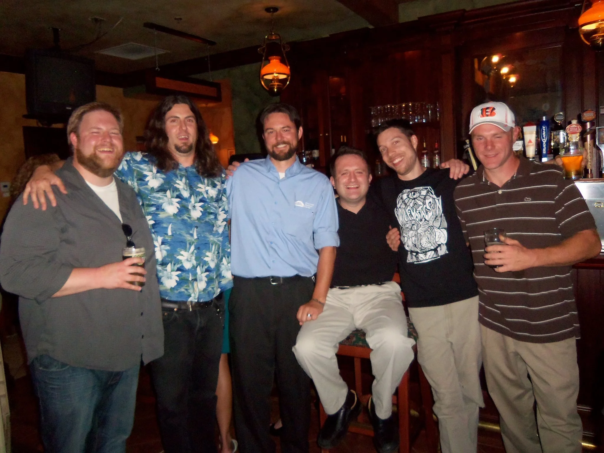Six men are in a bar, smiling and posing for a photo. One man is holding a drink, and others have their arms around each other's shoulders. They appear to be enjoying a night out.