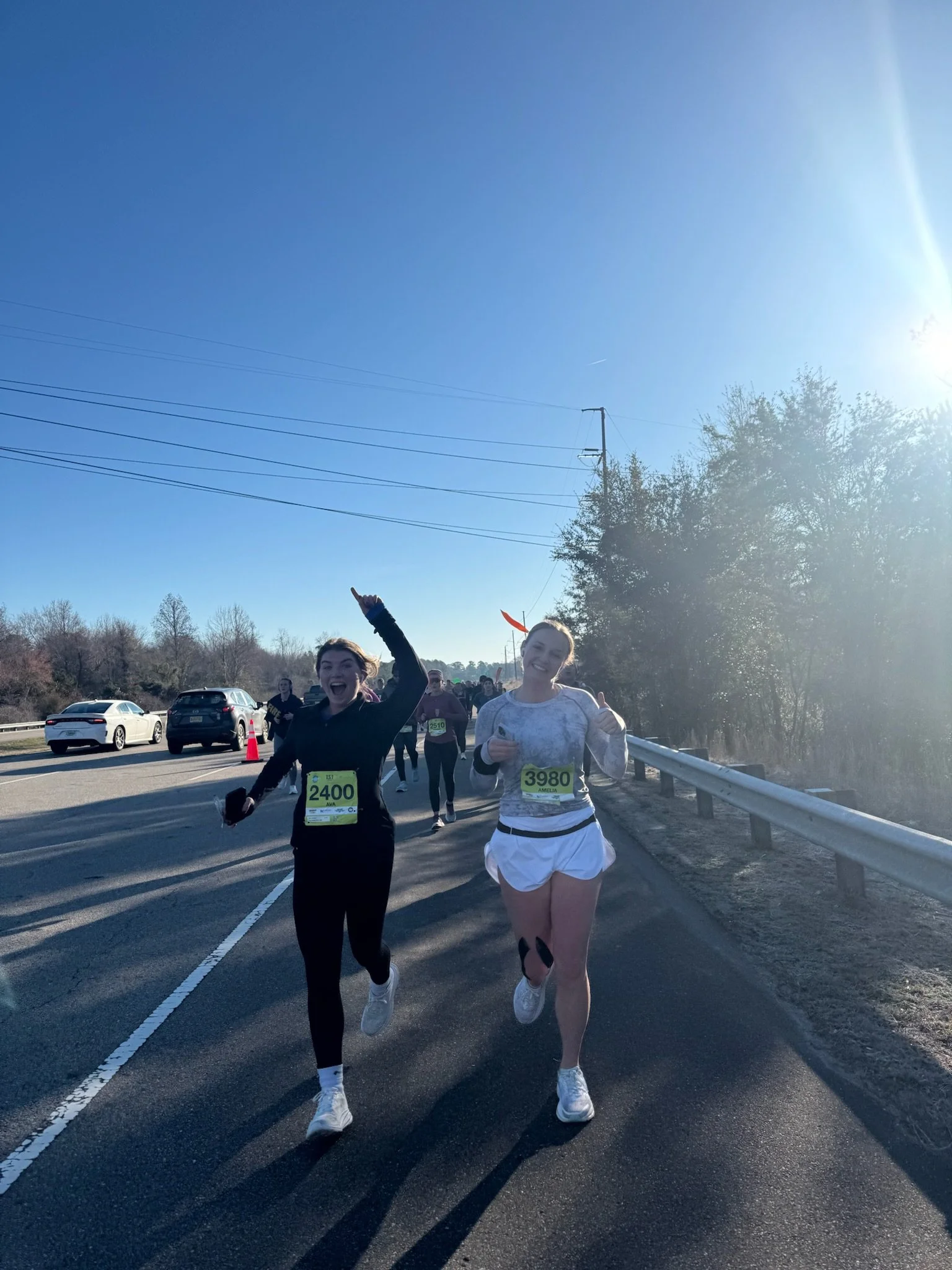 Two women running in a marathon or race on a paved road with traffic cones, other runners, parked cars, trees, and power lines in the background during daytime with clear blue sky.