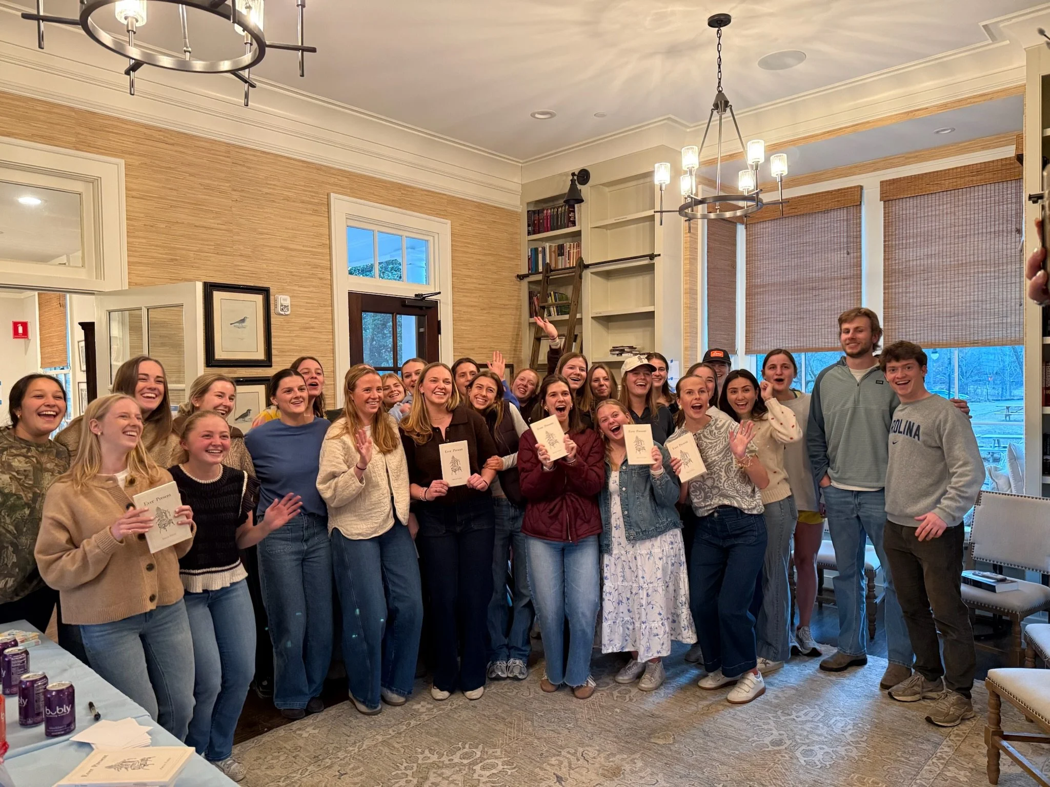 A group of young adults, mostly women, smiling and posing together indoors for a photo during a celebration. Some are holding books, and there is a table with purple cans and papers in the foreground. The room has large windows, bookshelves, and warm lighting.