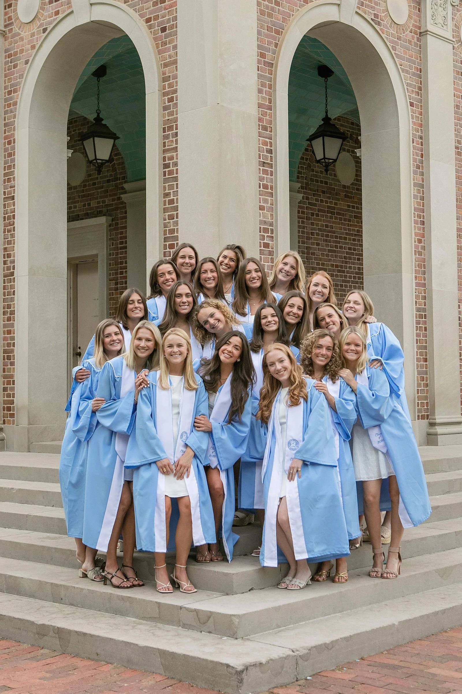 Group of women wearing light blue and white graduation gowns standing on steps outside a brick building with arches and lanterns.