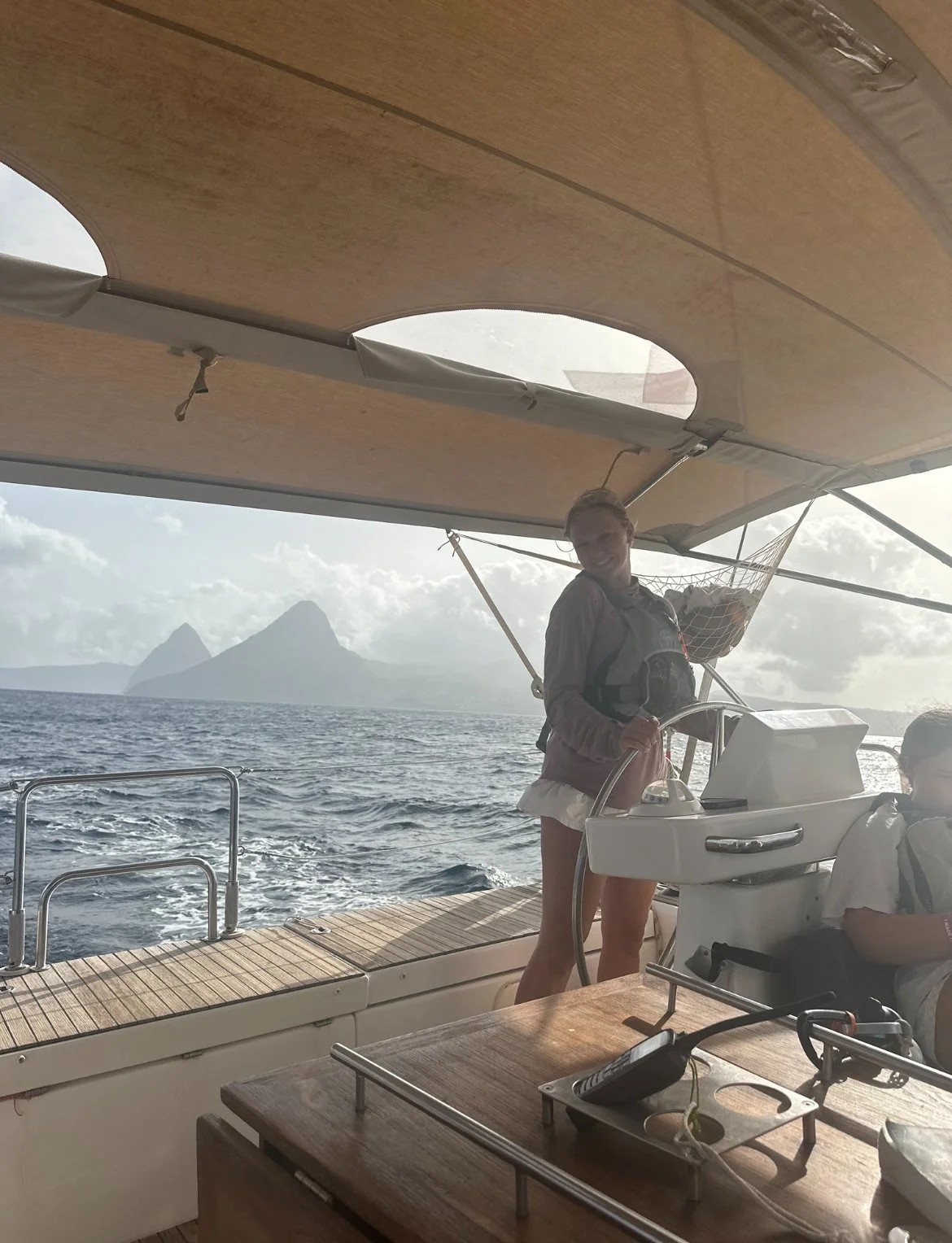 Smiling woman steering a boat on the water with an island in the background, a man seated nearby, and the boat's deck and controls visible.