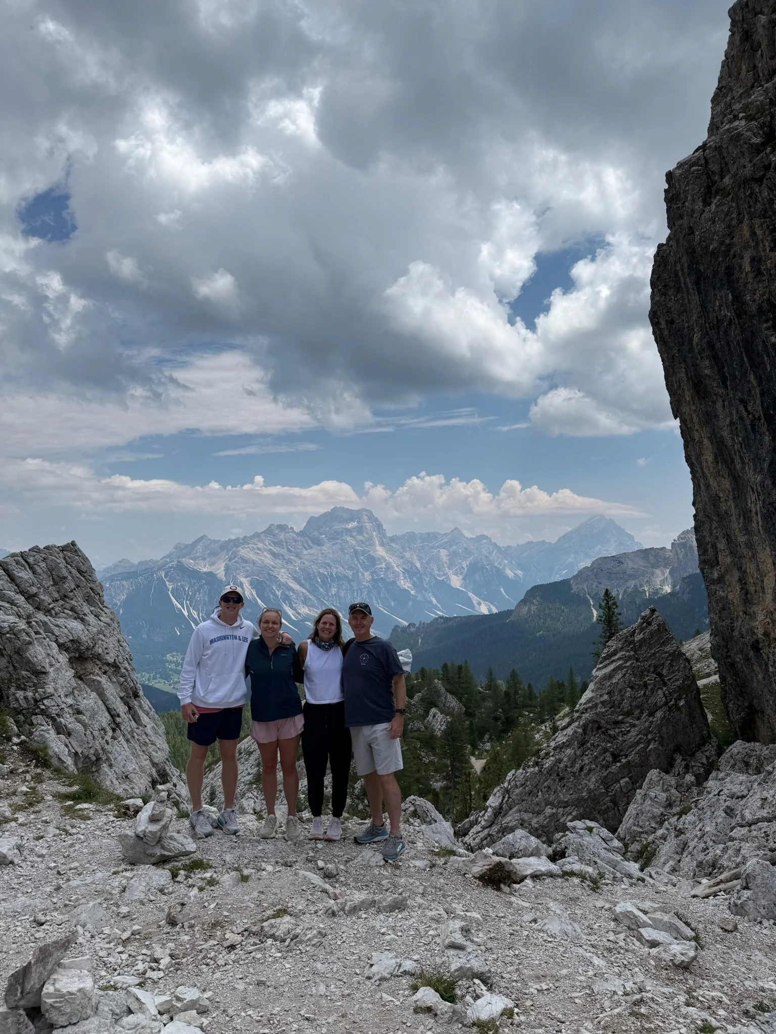 Group of four people standing on rocky mountain trail with mountain range and sky with clouds in the background.