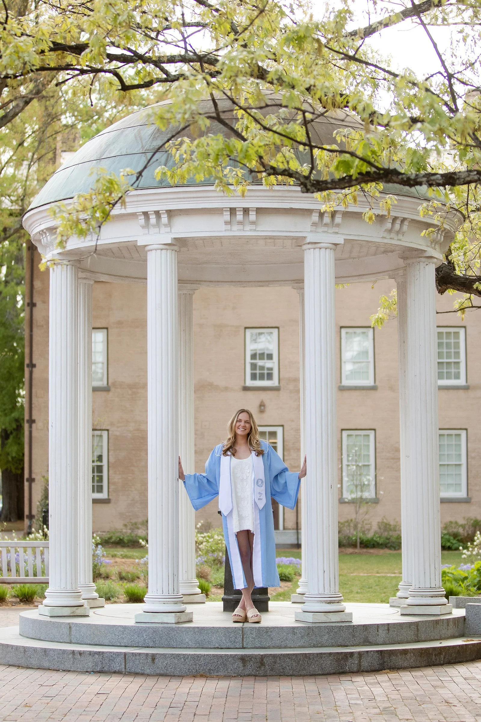 A joyful woman in a blue graduation gown and cap standing on the steps of a small white circular colonnade with her arms outstretched, celebrating her graduation outdoors.