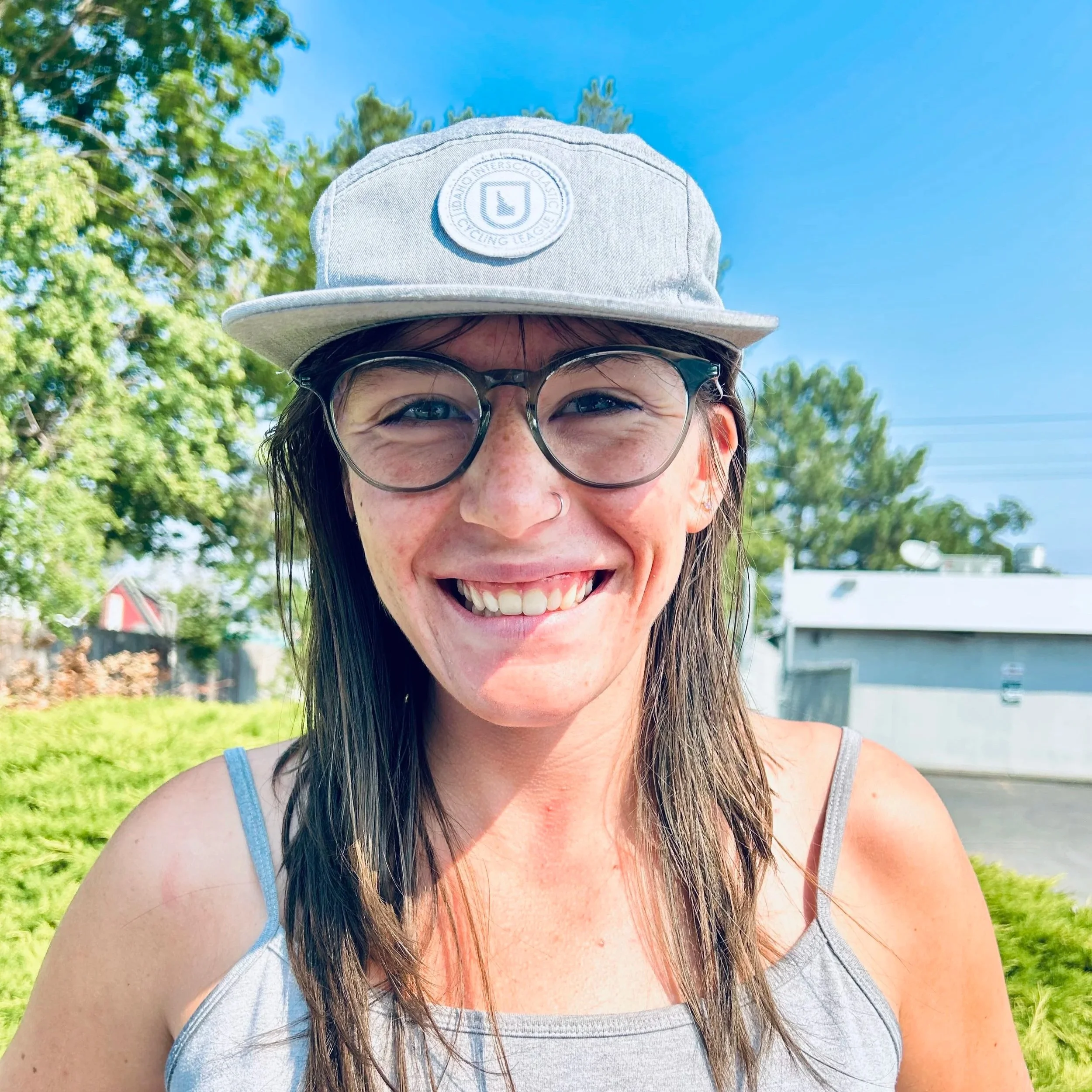 A smiling woman with freckles and glasses wearing a gray tank top and a light gray baseball cap standing outdoors with green trees and blue sky in the background.