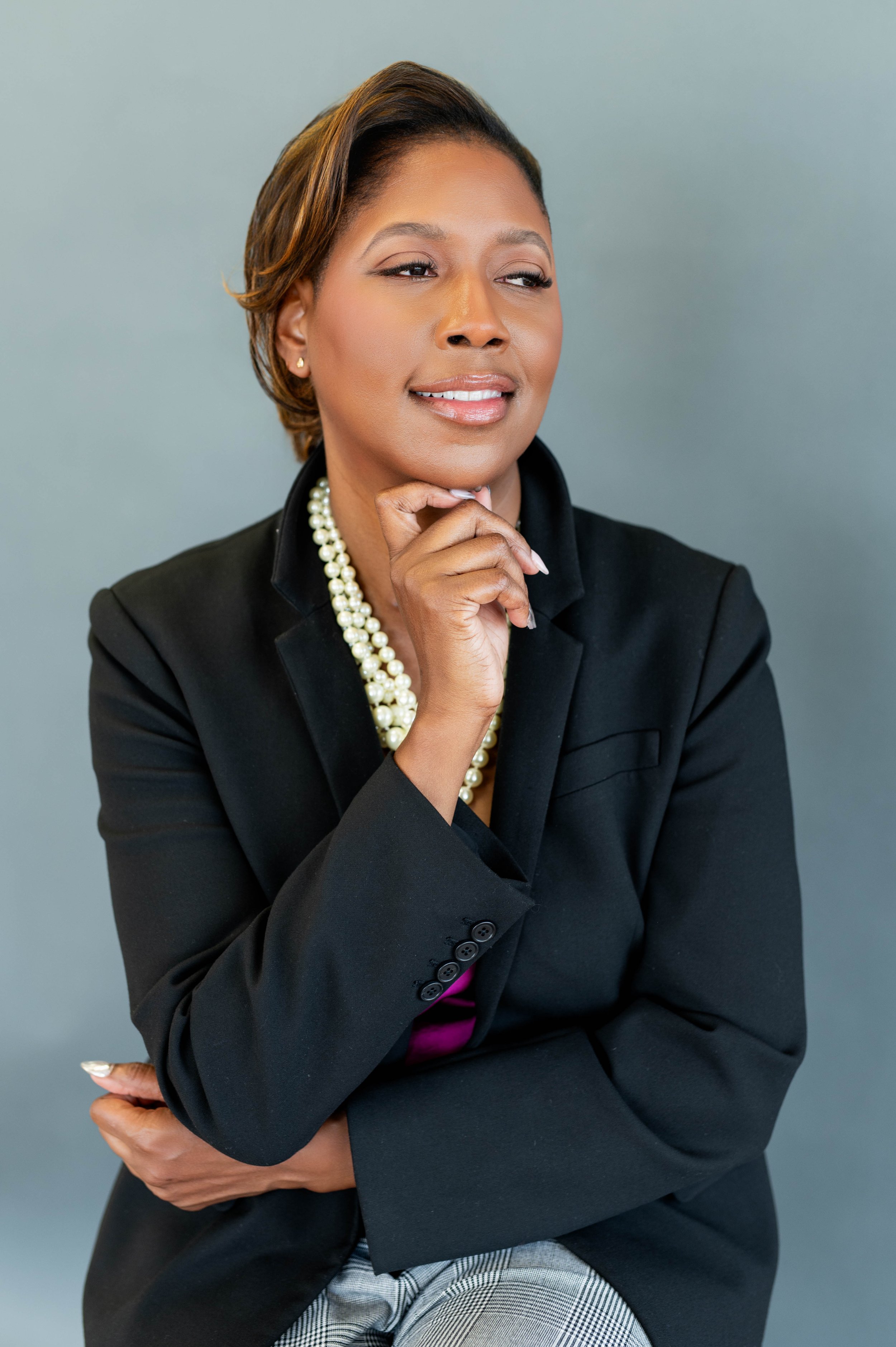 A woman with short hair wearing a black blazer, pearl necklace, and checkered pants, sitting against a plain gray background.
