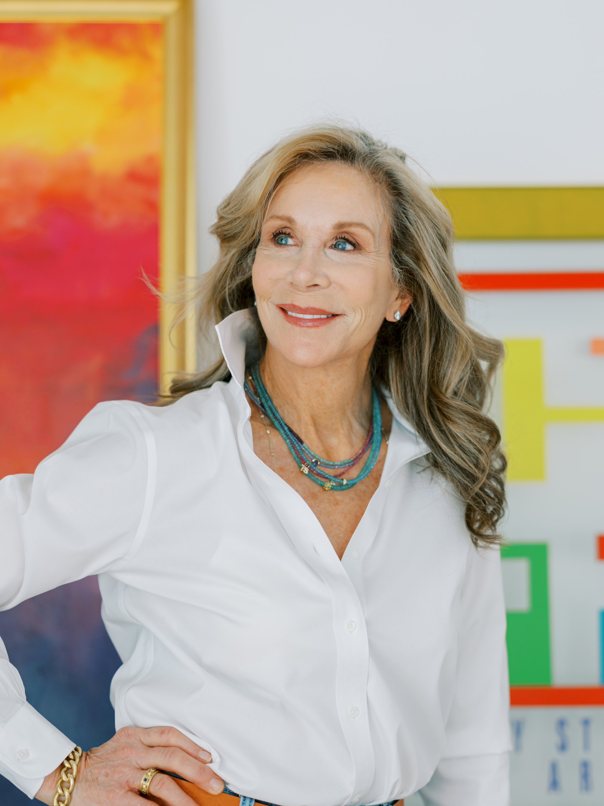 A smiling  woman with long, wavy blonde hair and blue eyes, wearing a white shirt, multi-colored necklaces, and jewelry, standing in front of an abstract colorful painting and chart.