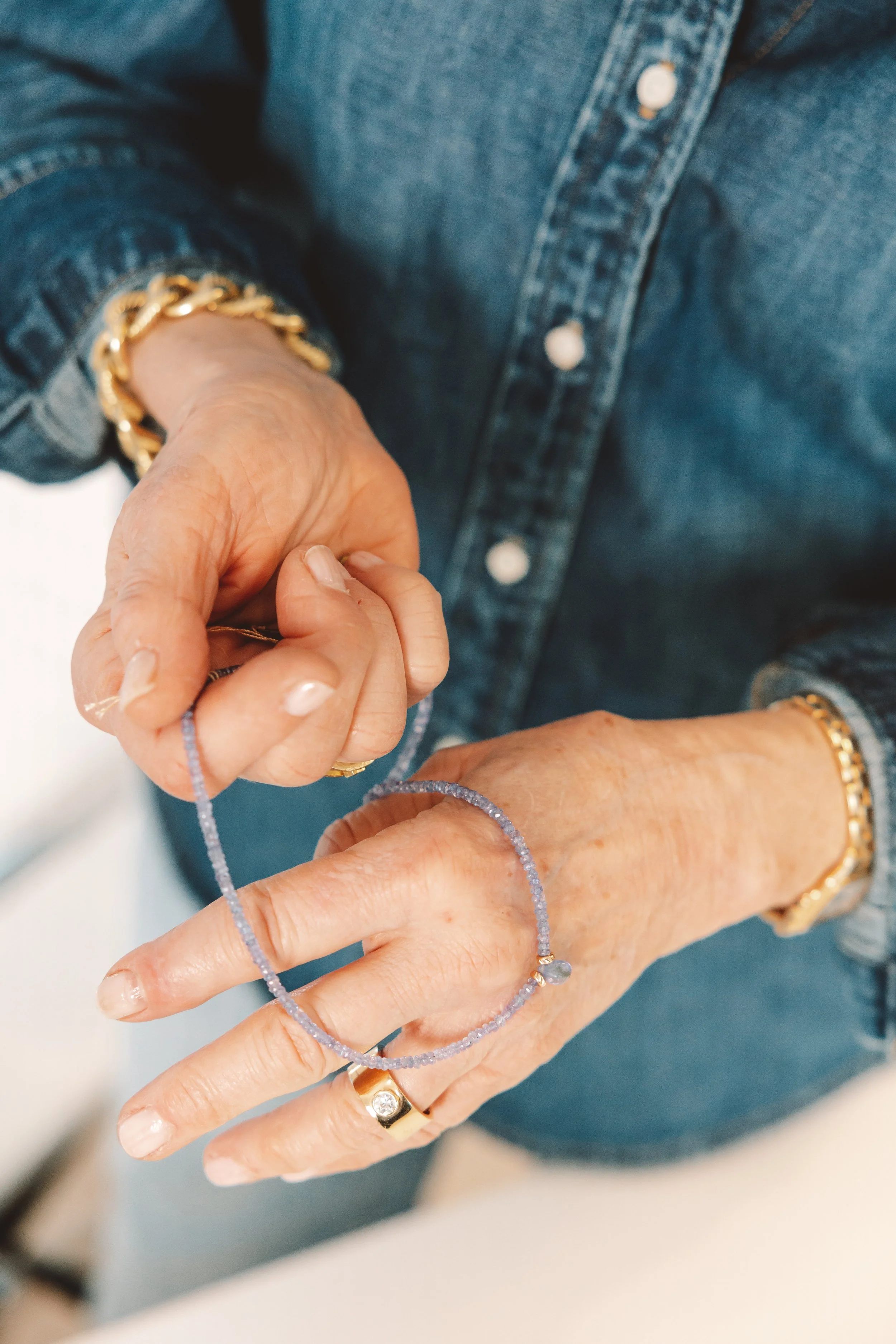 Close-up of a woman in a denim jacket and multiple gold rings and bracelets, holding a delicate light purple beaded bracelet.