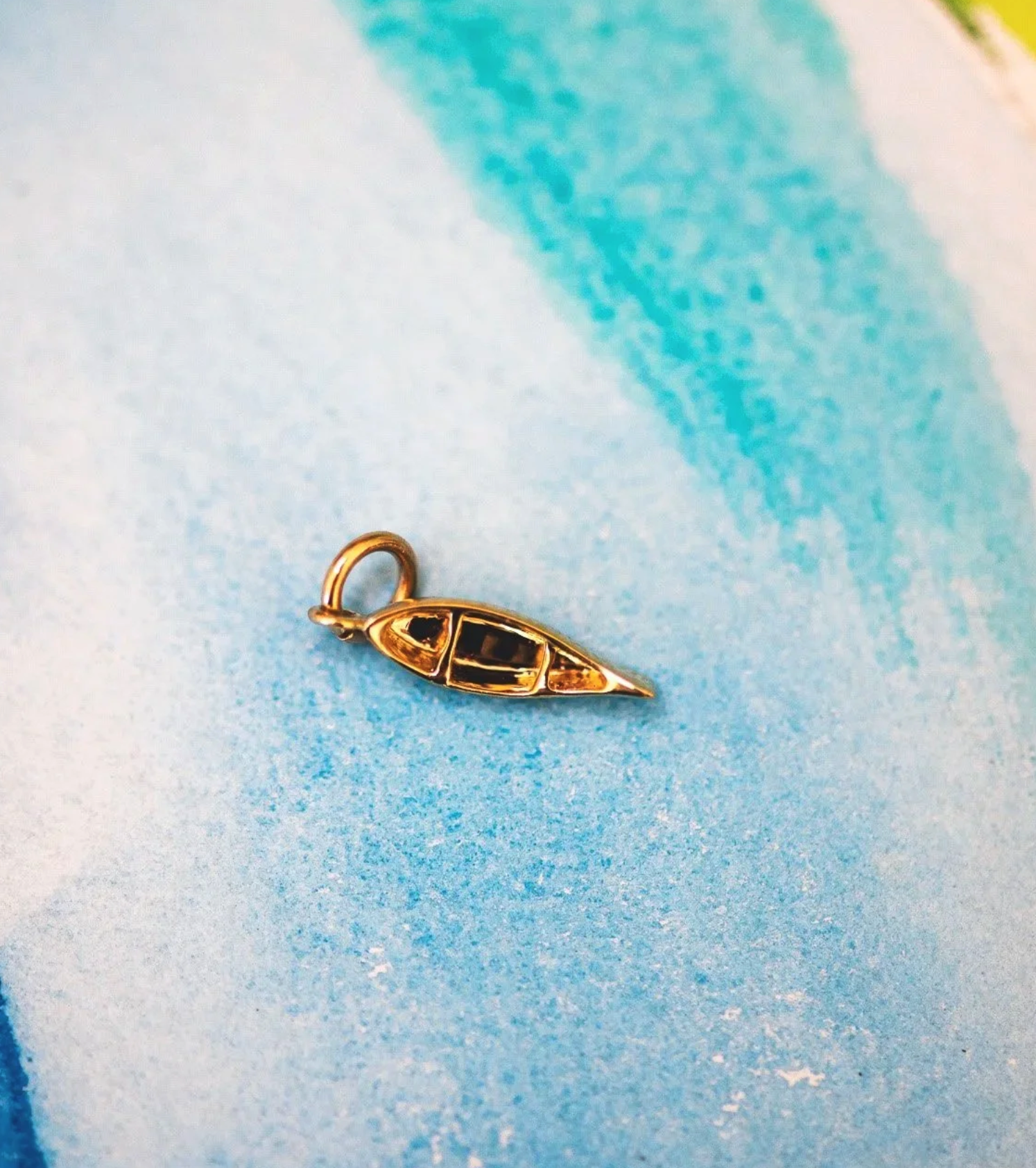 A small gold charm shaped like a canoe with black details, lying on a light blue textured surface.