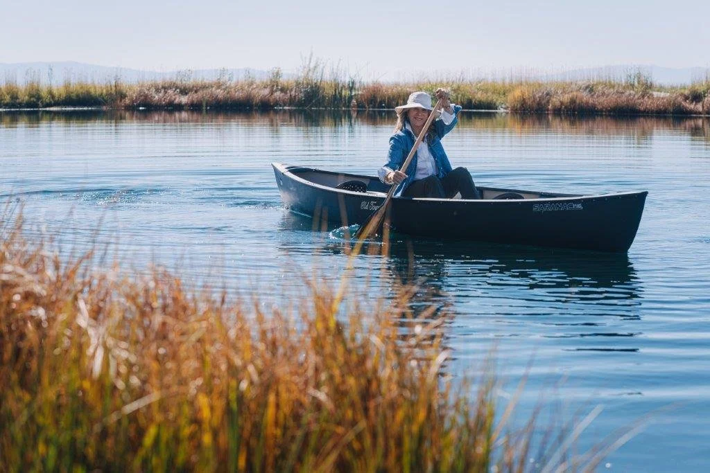 A woman in a hat and denim jacket paddles a canoe on a calm lake near tall grasses under a clear sky.