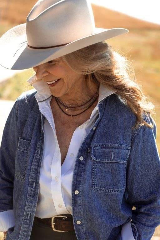 A smiling woman wearing a cowboy hat, denim jacket, white shirt, and layered necklaces outdoors.