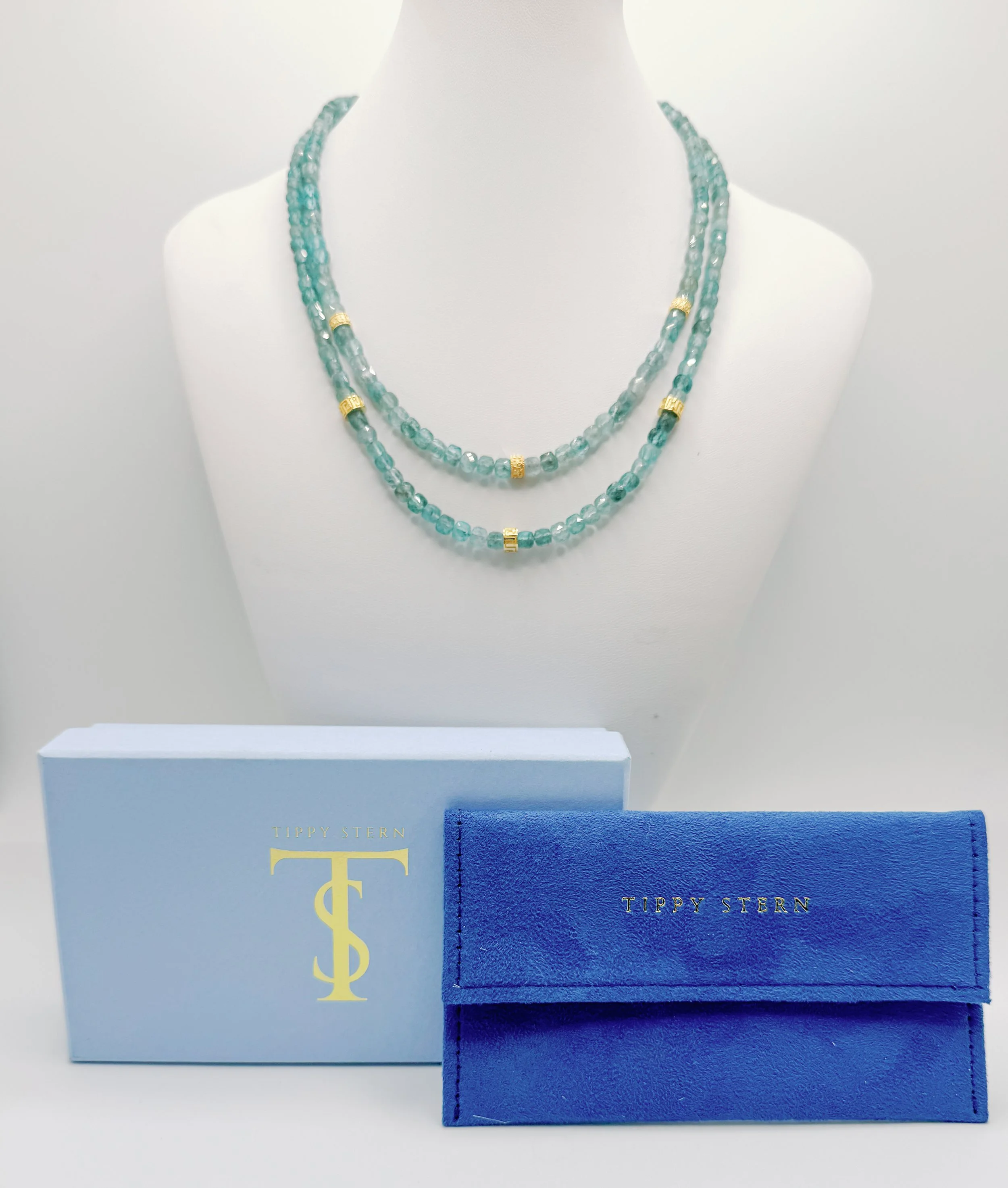 Two strands of aquamarine beads with gold accents draped on a white display bust, alongside a blue jewelry box and a partial view of a dark blue box with gold lettering.