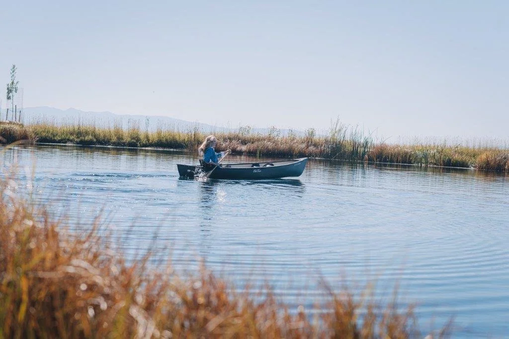 A woman in a blue jacket paddles a canoe on a calm river surrounded by tall grass and reeds, with mountains in the background on a clear day.