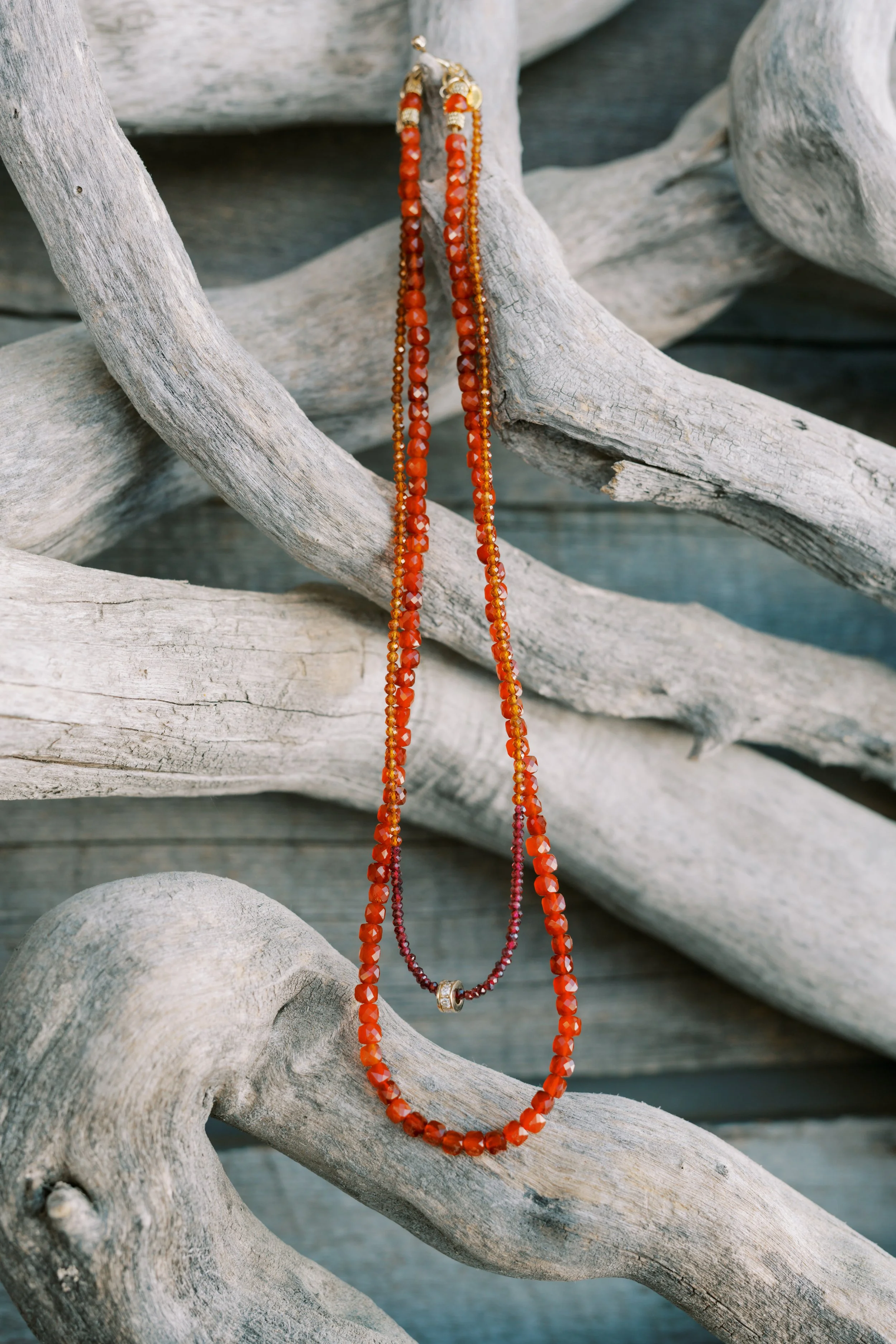A necklace with multiple strands of small, faceted orange and red beads, featuring two gold and bead accents, hanging over weathered driftwood.