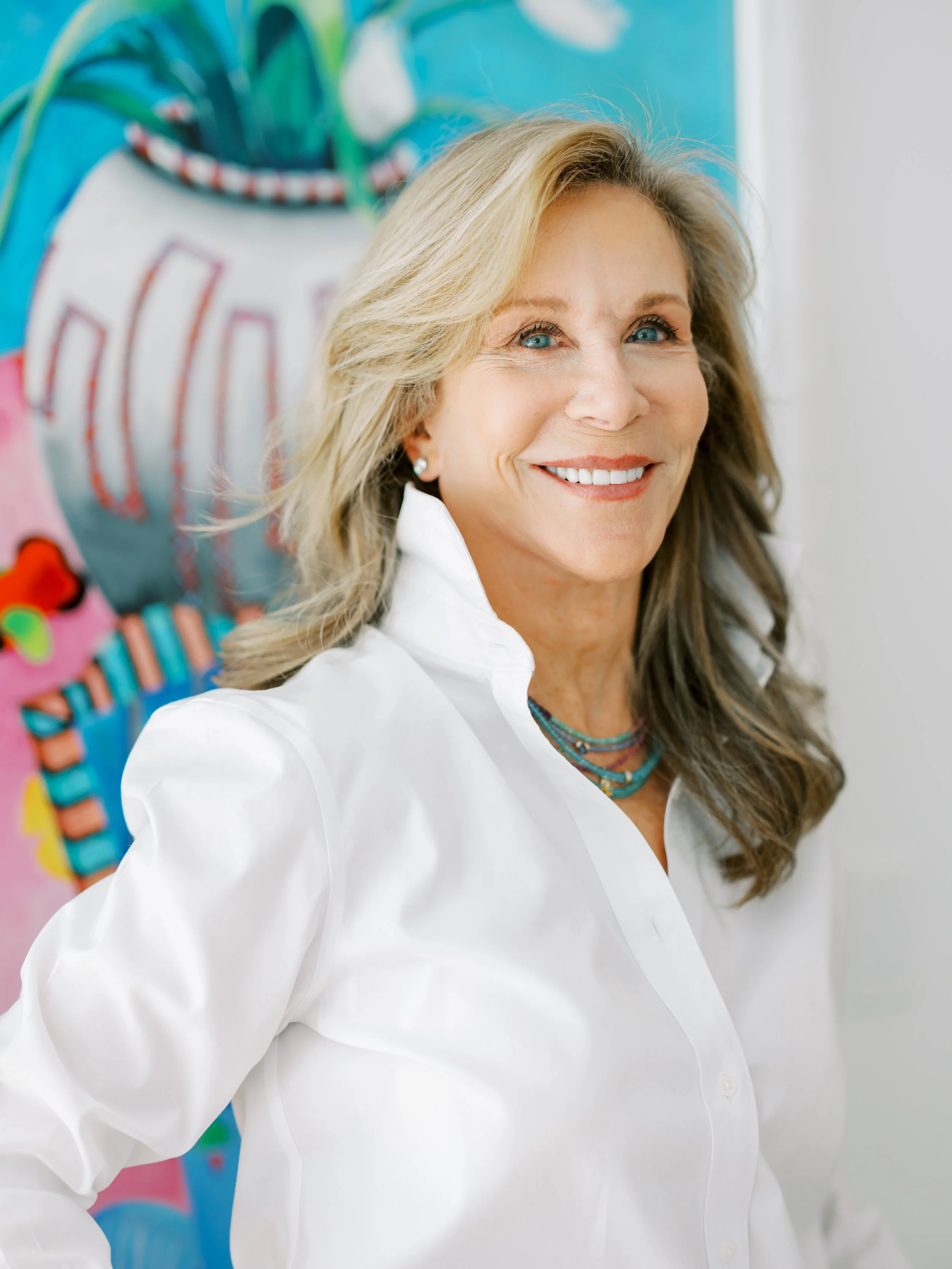 A woman with blonde hair and blue eyes smiling, wearing a white shirt and a colorful beaded necklace, standing in front of colorful art. 