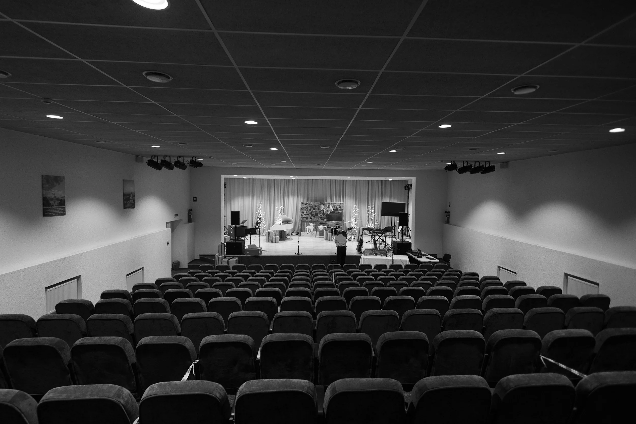 An auditorium or theater with rows of empty chairs facing a stage decorated with holiday-themed props, including a Christmas tree and presents, and a person setting up or adjusting equipment on the stage.