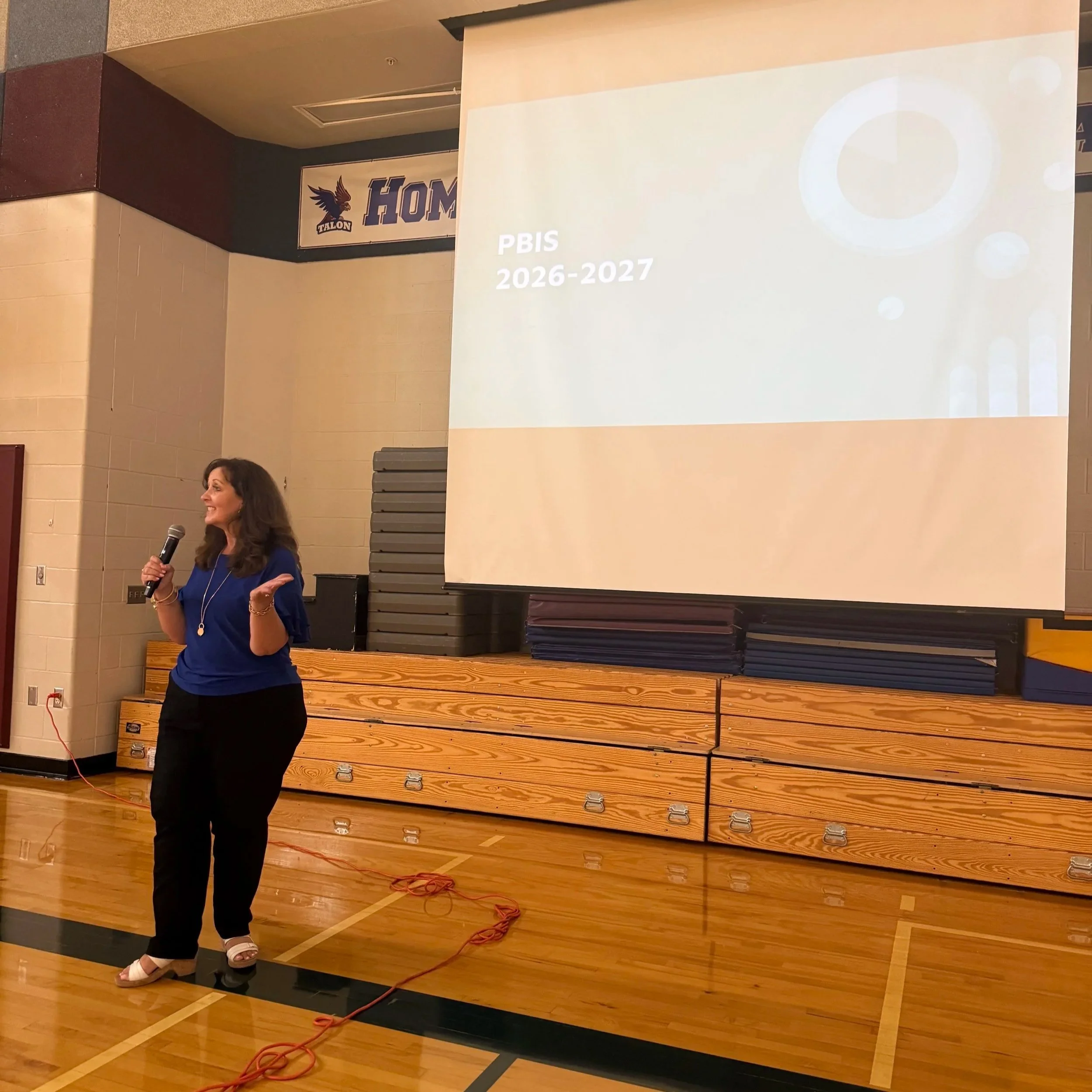A woman with long brown hair, wearing a blue top and black pants, is speaking into a microphone in a gymnasium. Behind her is a large white projection screen displaying the text "PBIS 2026-2027." To the left of the screen is a stacked set of gym mats. A banner with a hawk mascot and the word "TALON" is hung on the wall in the background.