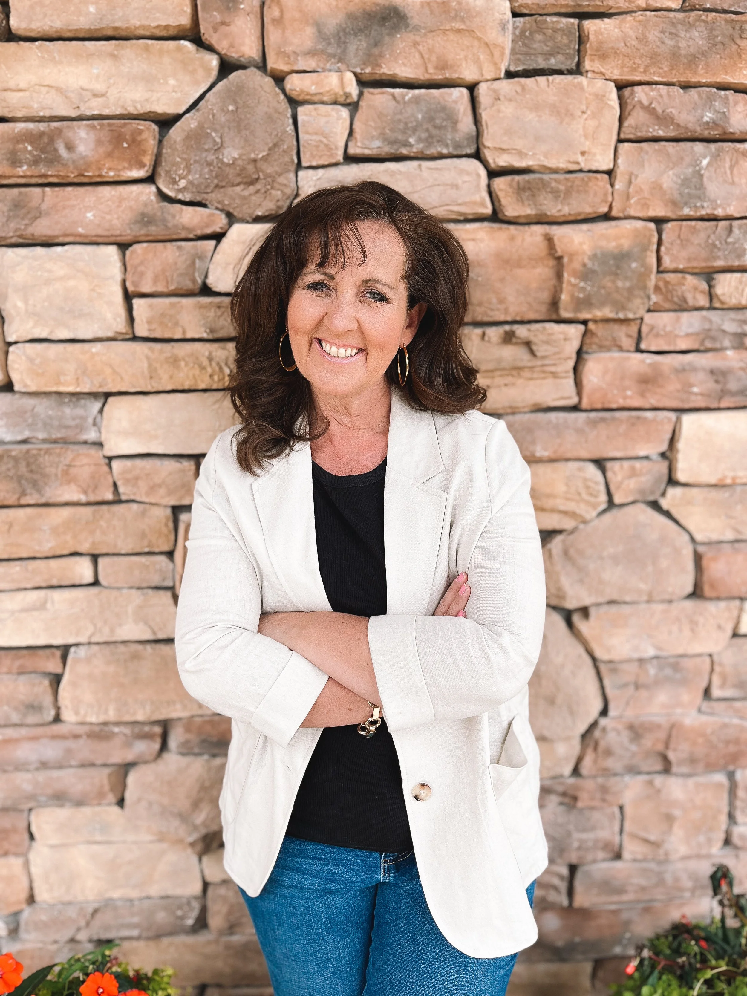 A woman with brown hair, wearing hoop earrings, a black top, a light-colored blazer, and blue jeans, stands with her arms crossed in front of a stone wall. She is smiling.
