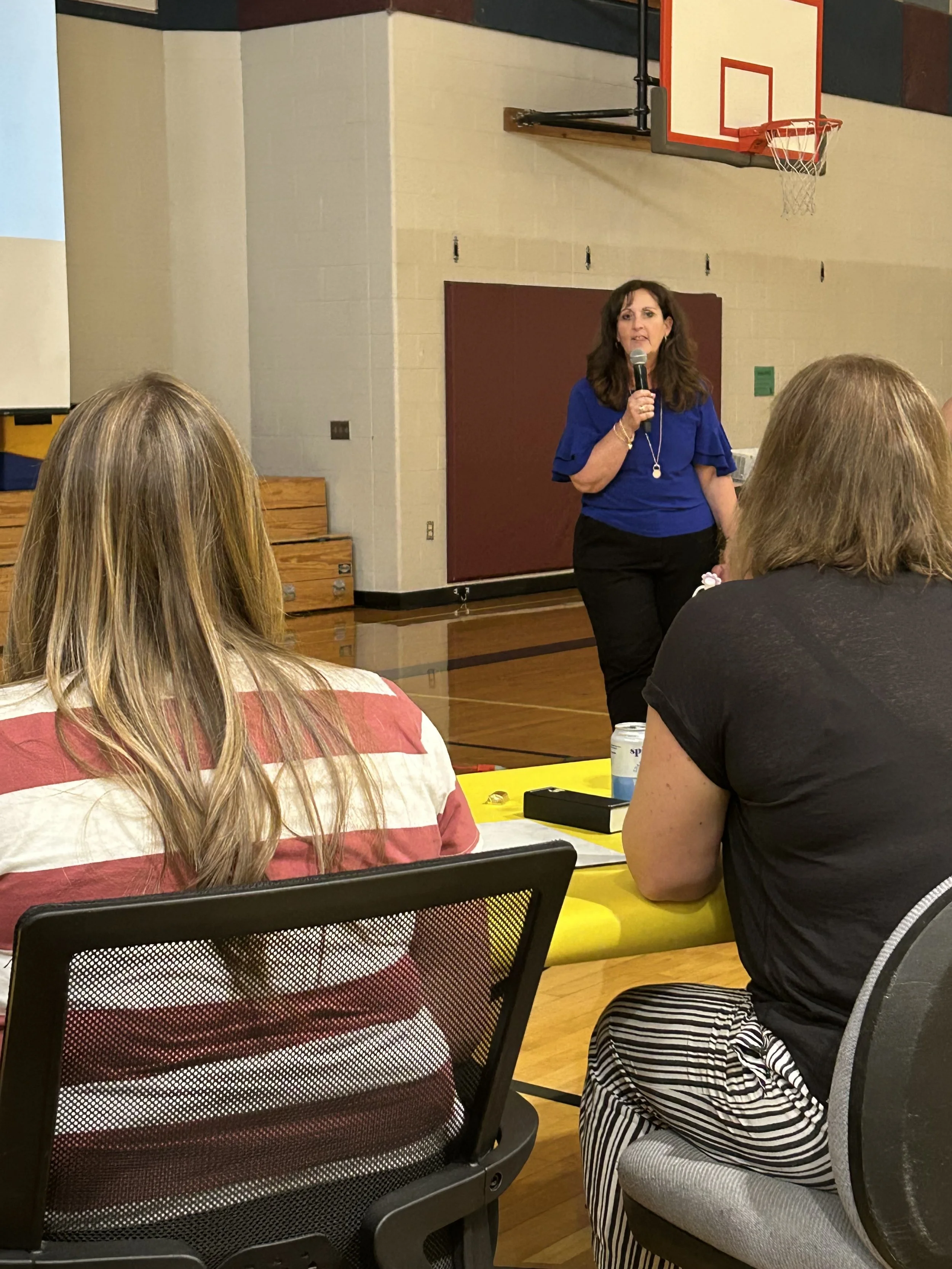 A woman in a blue shirt is speaking into a microphone during a presentation or meeting in a gymnasium or multi-purpose room. There are audience members seated at tables, and a basketball hoop is visible on the wall behind her.