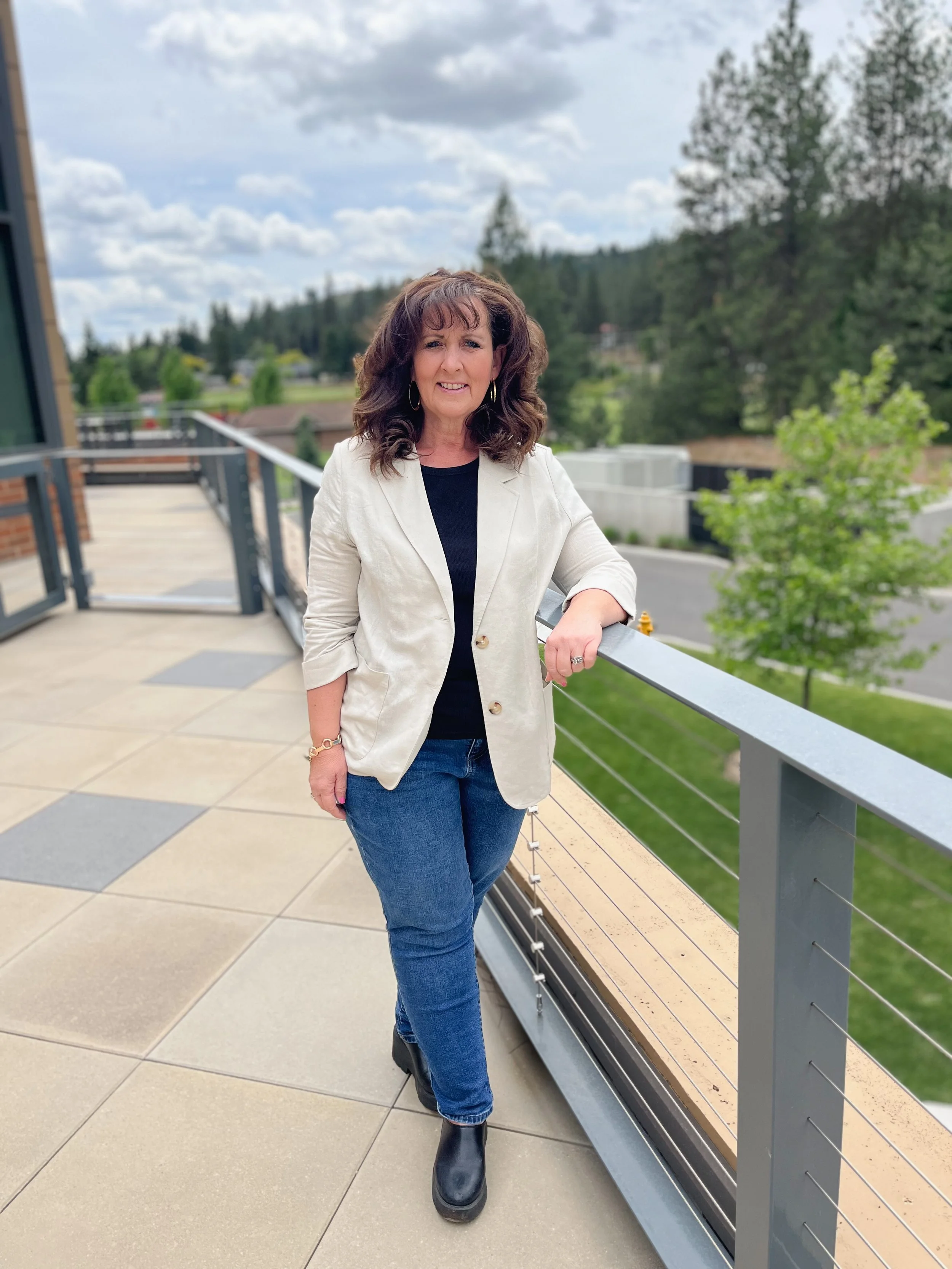 A woman with brown hair and hoop earrings standing on a balcony with a view of trees and a cloudy sky, wearing a white blazer, black top, blue jeans, and black boots, smiling and leaning on a metal railing.