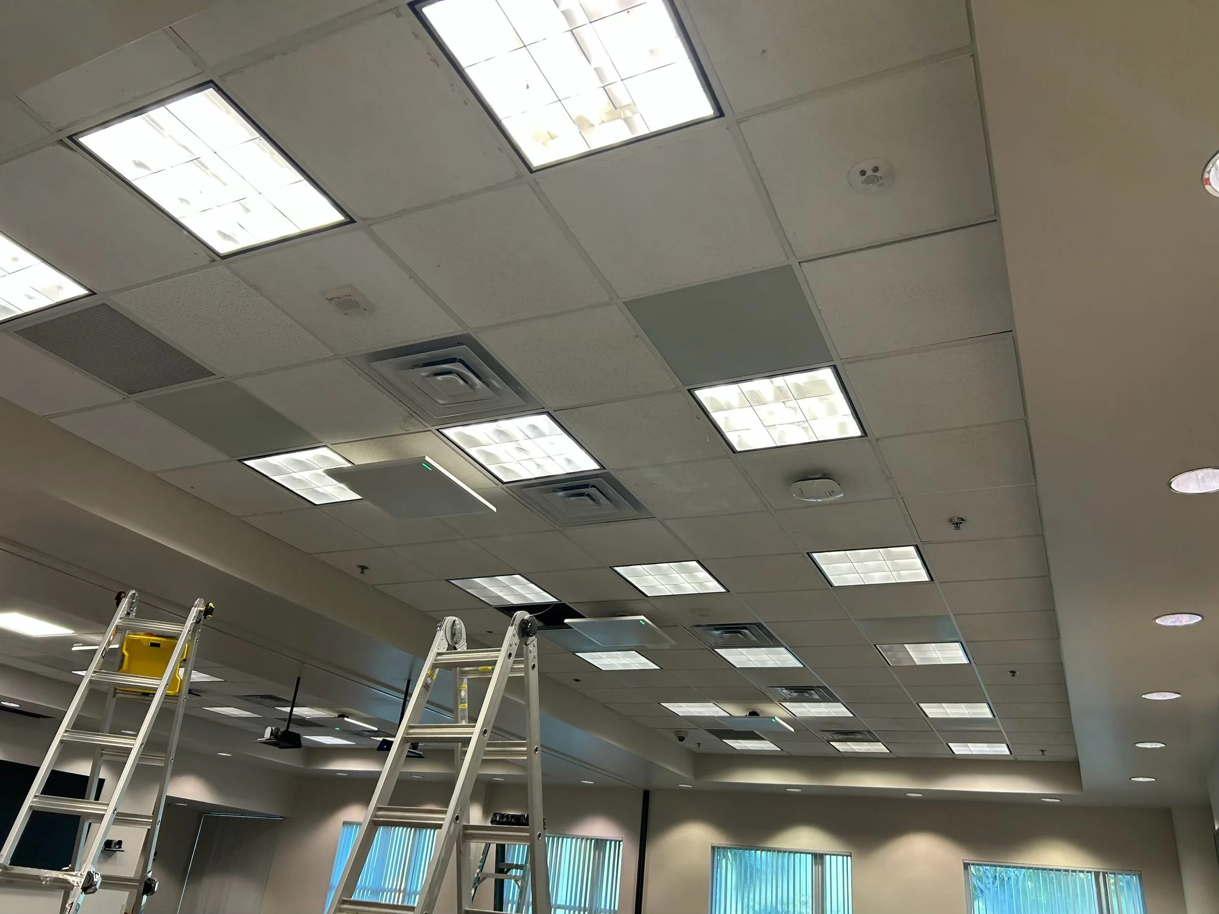 Ceiling with fluorescent lights, air vents, and smoke detector, partial view of ladders, inside a building.