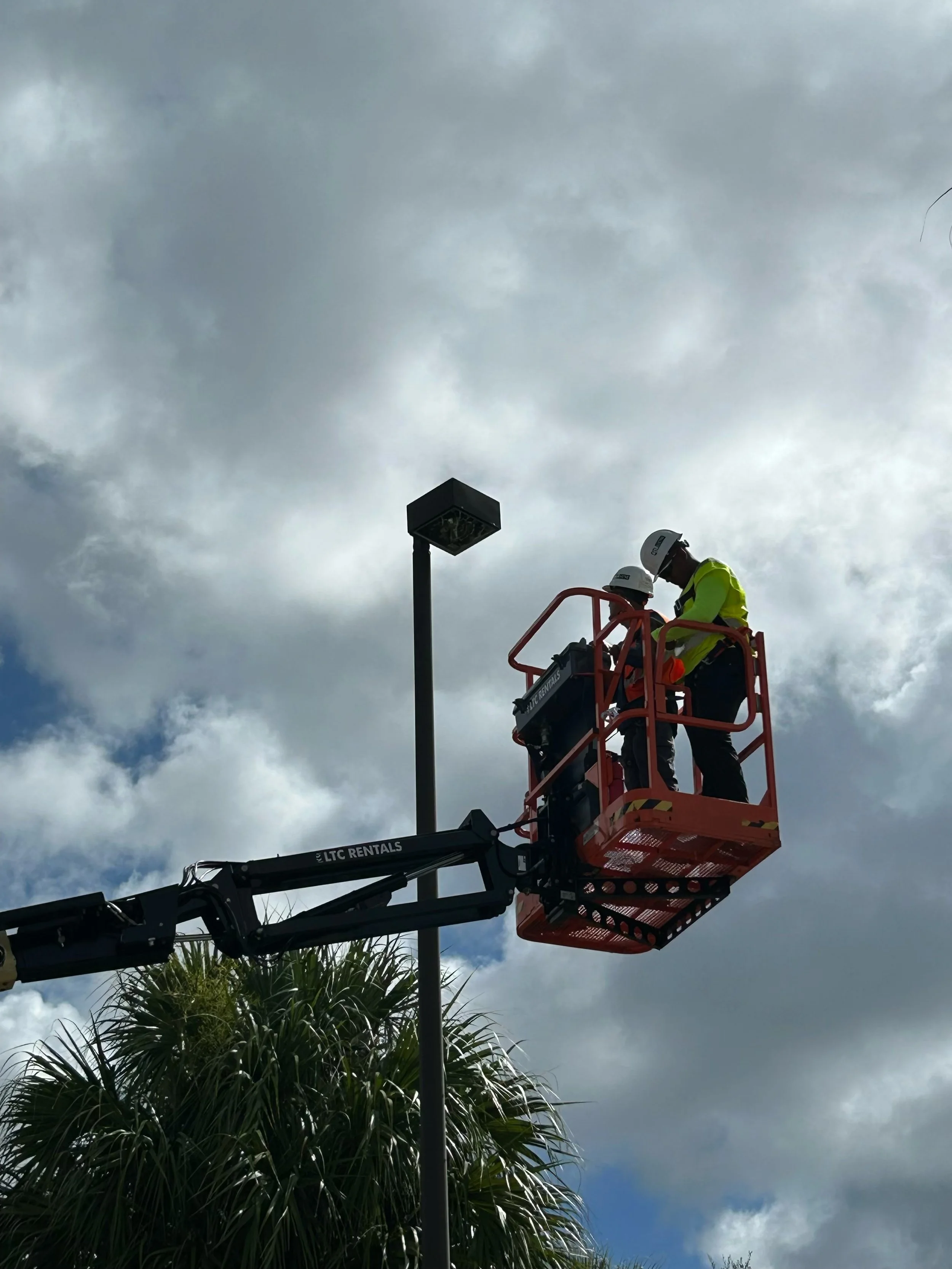 Two workers in safety helmets and high-visibility vests are using a lift truck to reach a streetlight under a cloudy sky.